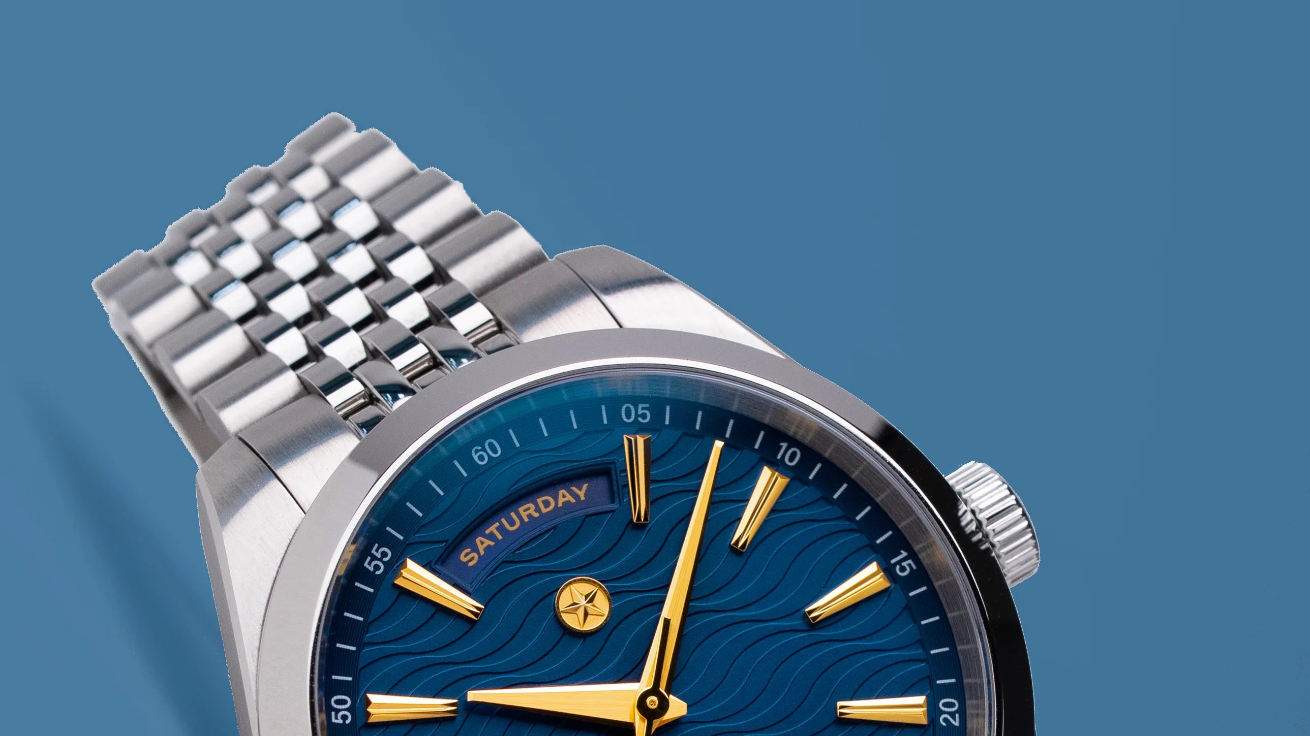 Close-up of a silver metal wristwatch with a blue textured dial, gold hour markers, and a day display showing "Saturday.