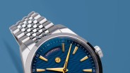 Close-up of a silver metal wristwatch with a blue textured dial, gold hour markers, and a day display showing "Saturday.