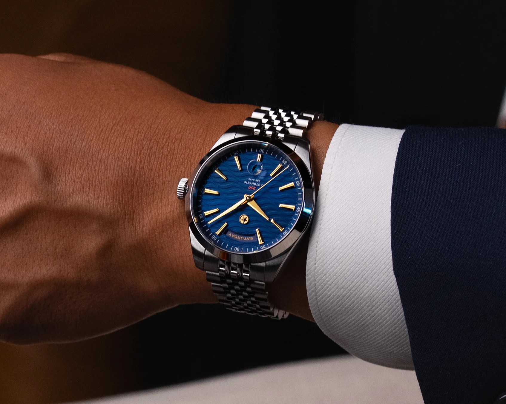 Silver metal wristwatch with blue textured dial and gold hour markers on a wrist with a white shirt cuff.
