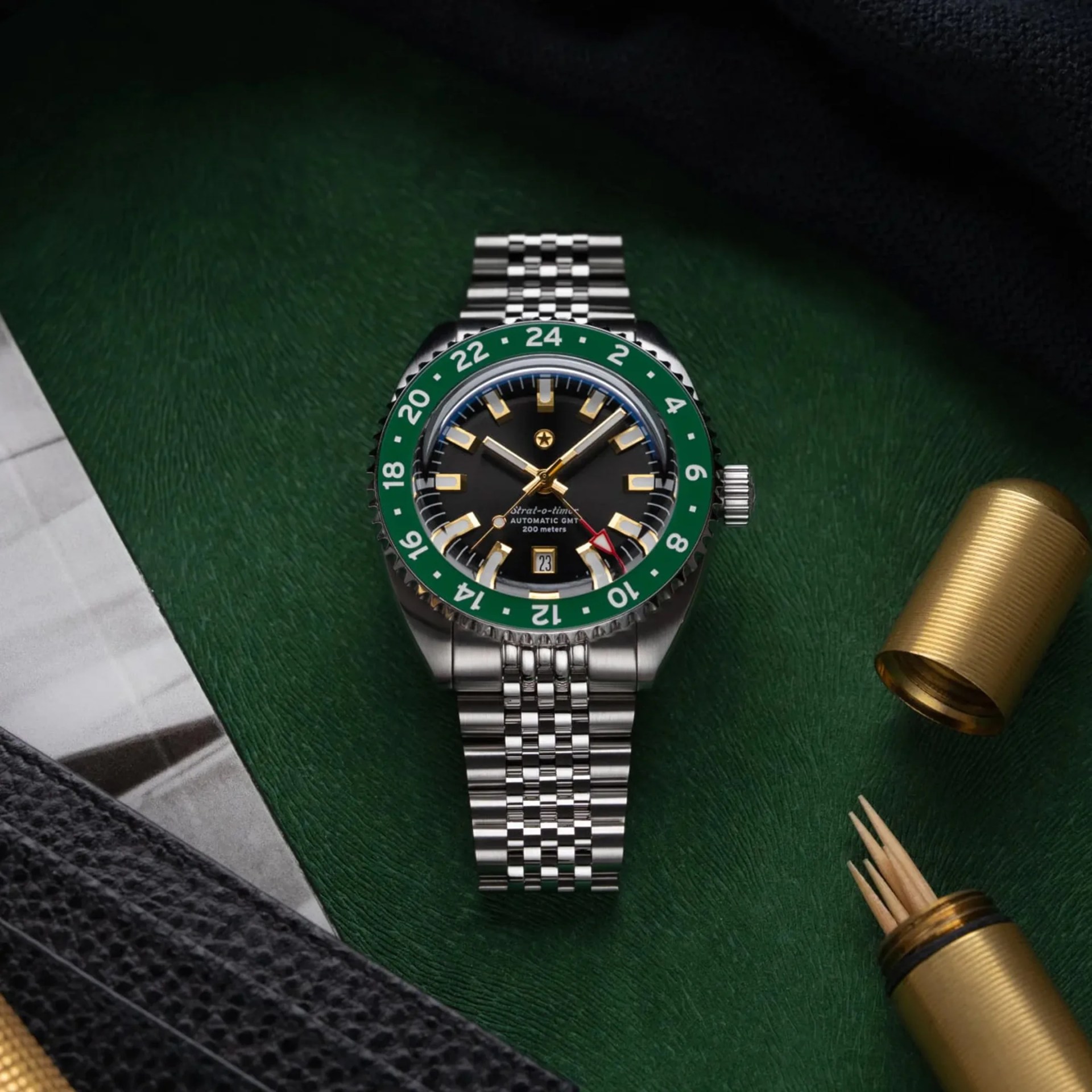 Stainless steel wristwatch with black dial, gold hour markers, green bezel, and metal link bracelet on green leather surface.