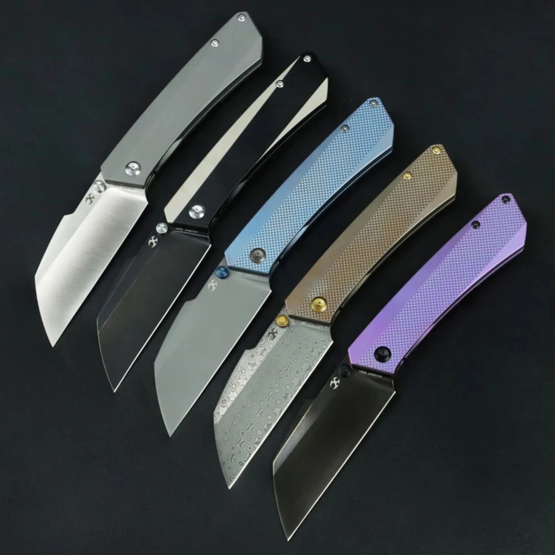 Five folding knives with various handle colors and blade finishes arranged diagonally on a black surface.