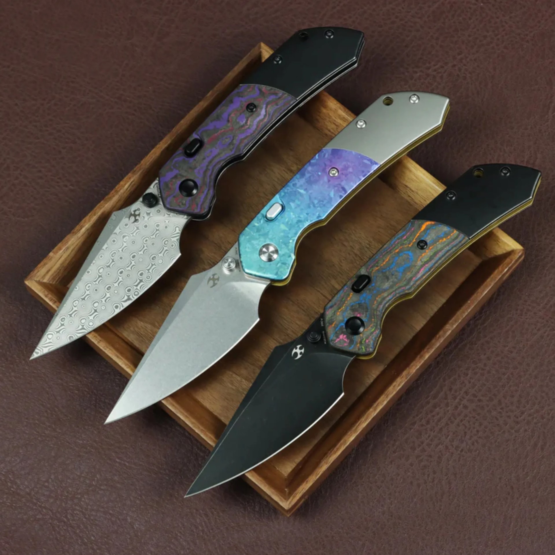 Three folding knives with patterned and metallic handles in a wooden tray on a brown leather surface.