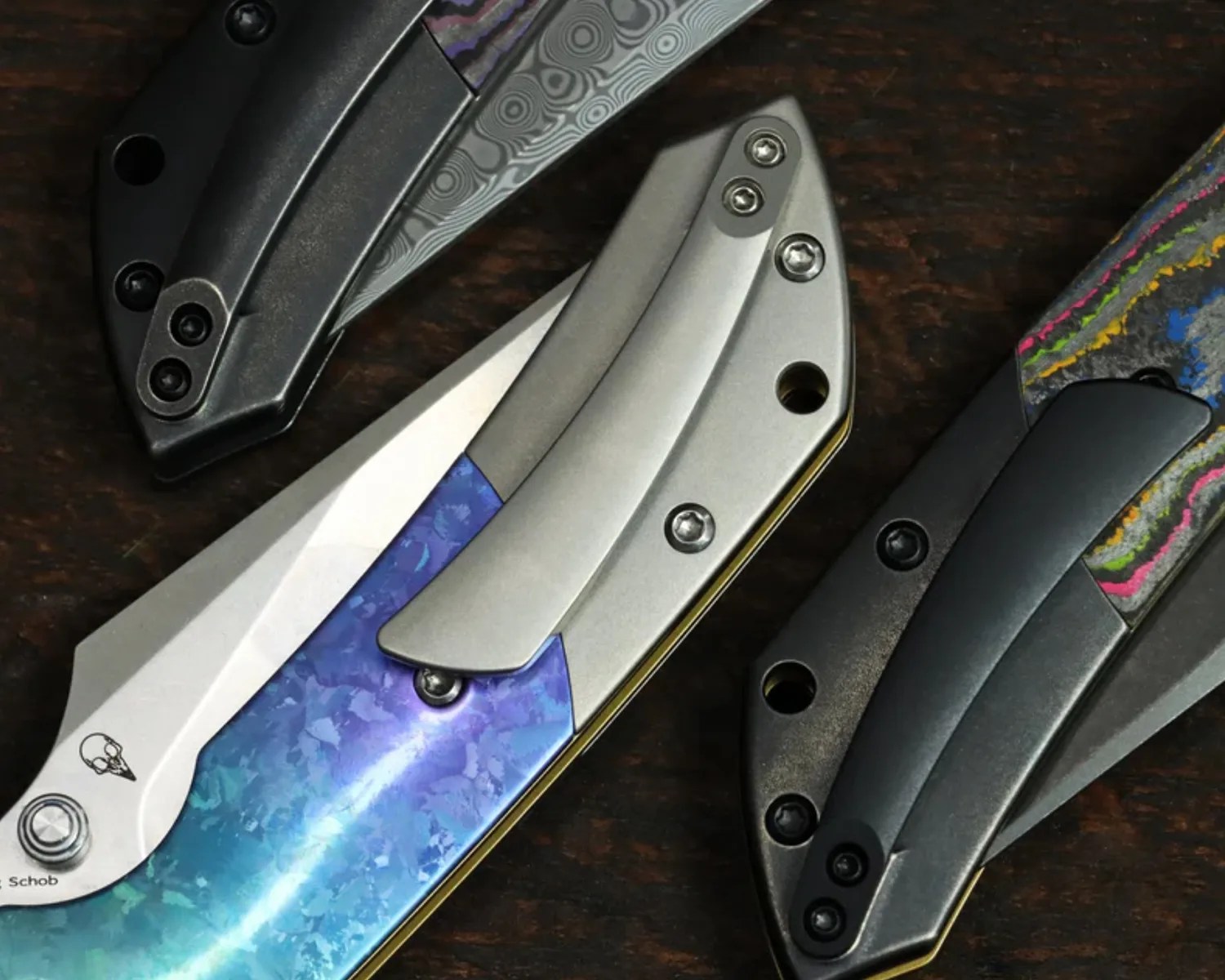 Three folding knives with colorful and patterned handles on a dark wooden surface.