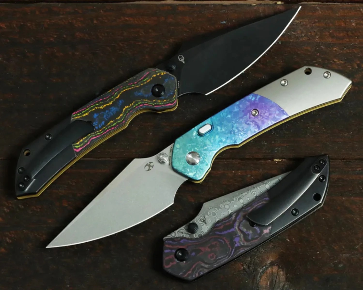 Three folding knives with colorful patterned handles and metal blades on a dark wooden surface.