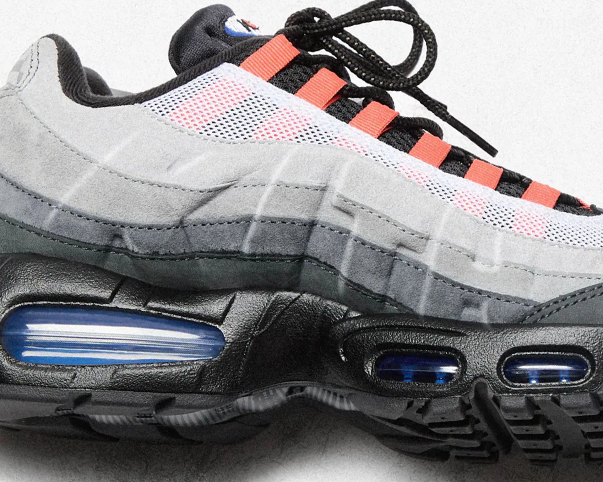 Black and gray sneaker with blue air cushioning and orange lace loops on a white background.