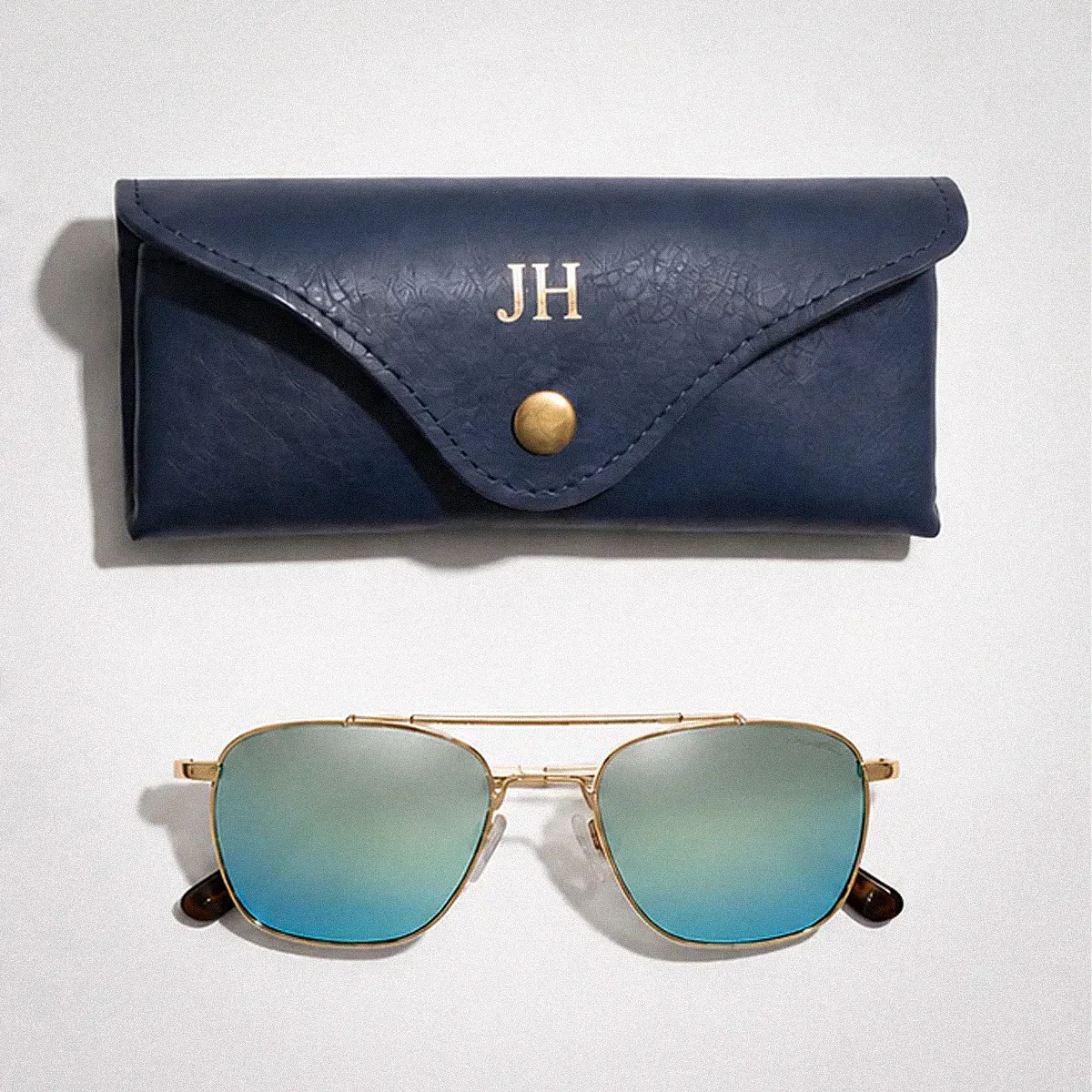 Blue leather sunglasses case with gold button and initials "JH" above gold-framed sunglasses with blue gradient lenses.