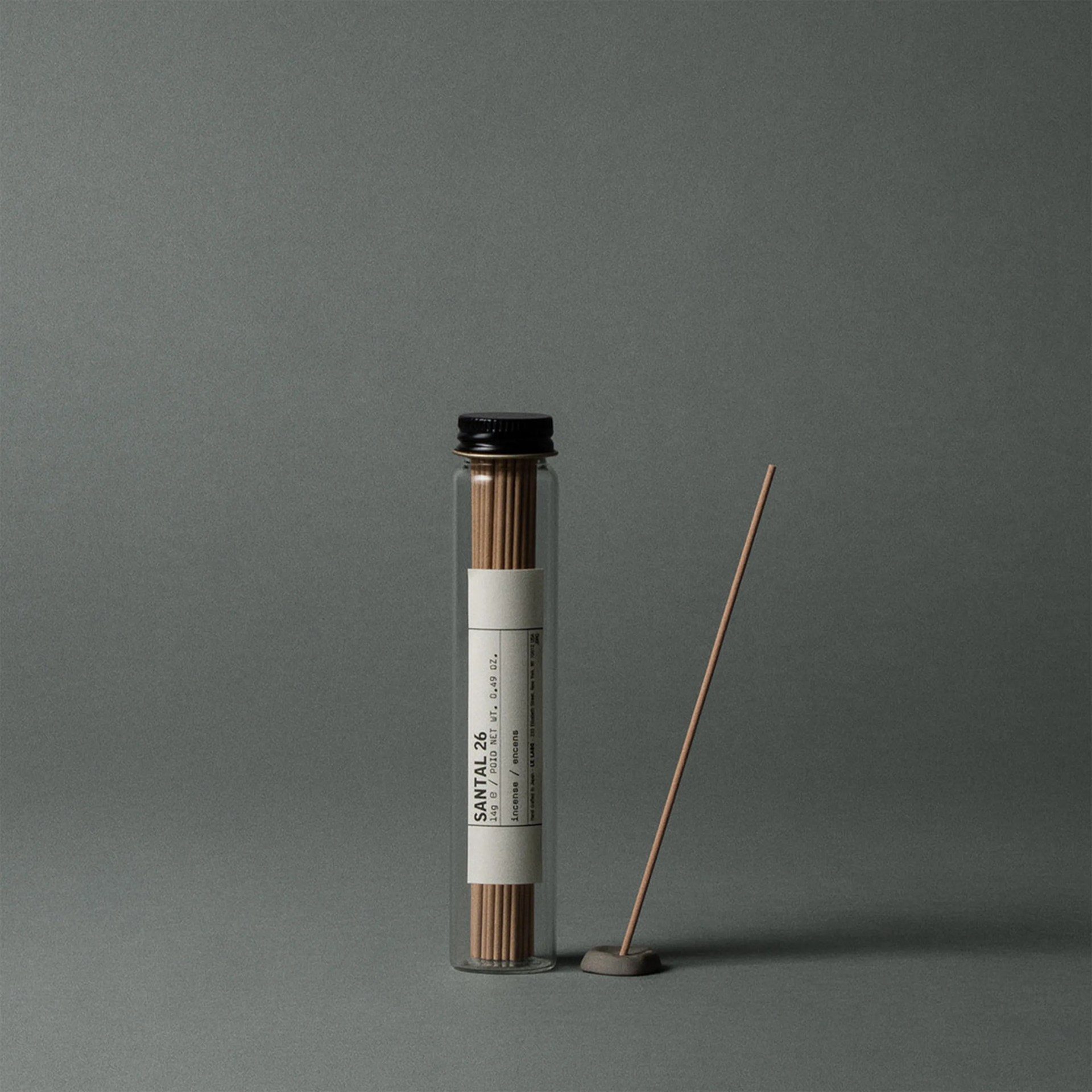 Clear glass tube with a black cap containing incense sticks labeled "SANTAL 26" next to a lit incense stick in a small gray holder.