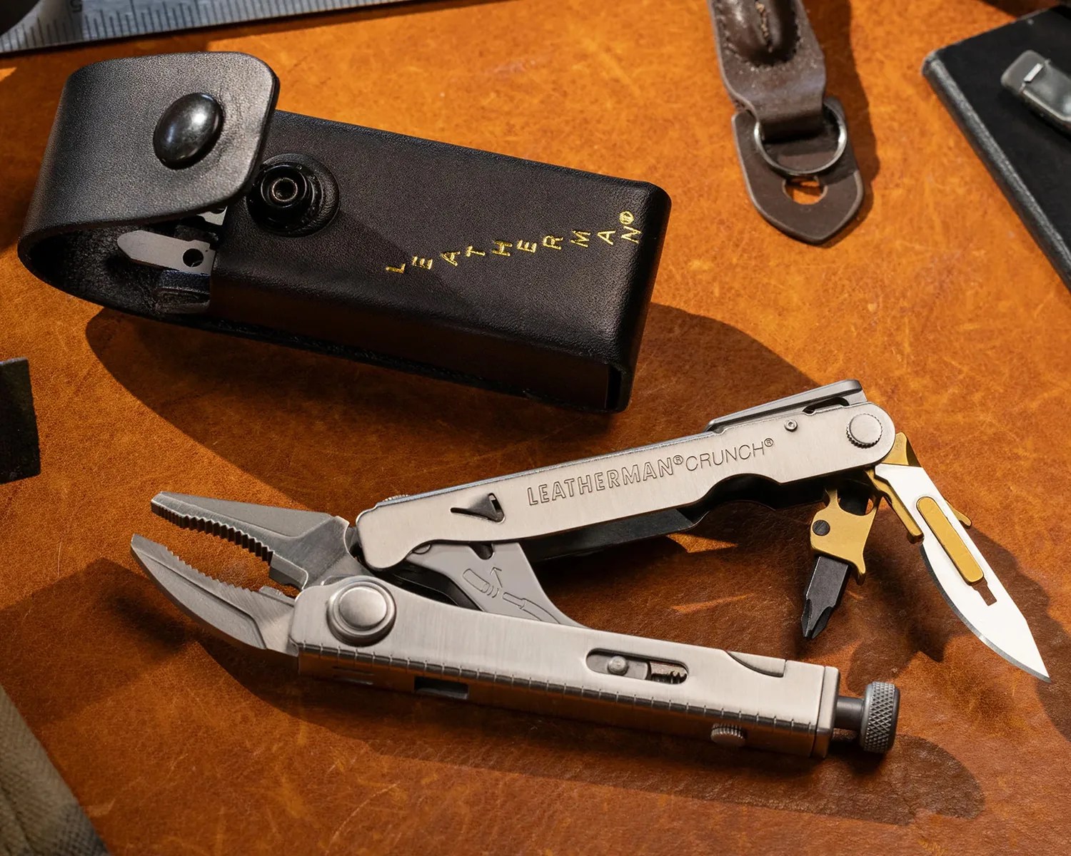 Stainless steel Leatherman Crunch multi-tool with pliers, knife, and screwdriver, next to a black leather sheath on a brown surface.