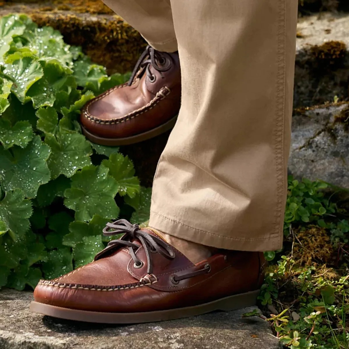 Brown leather boat shoes with dark laces worn with beige pants on a stone surface near green plants.