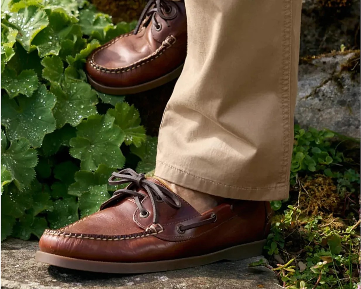 Brown leather boat shoes with dark laces worn with beige pants outdoors on stone and greenery.