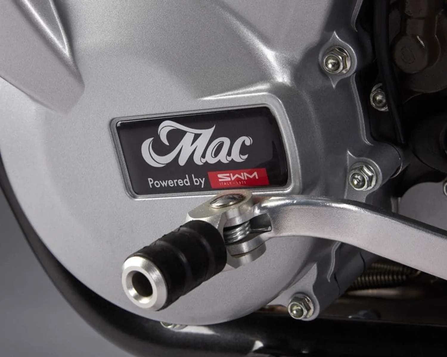 Close-up of a silver motorcycle engine cover with a "Mac Powered by SWM Italy 1971" label and a foot lever with a black rubber tip.