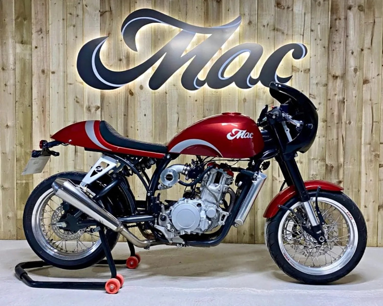 Red and black cafe racer motorcycle with silver exhaust and "Mac" logo on fuel tank, displayed on a stand.