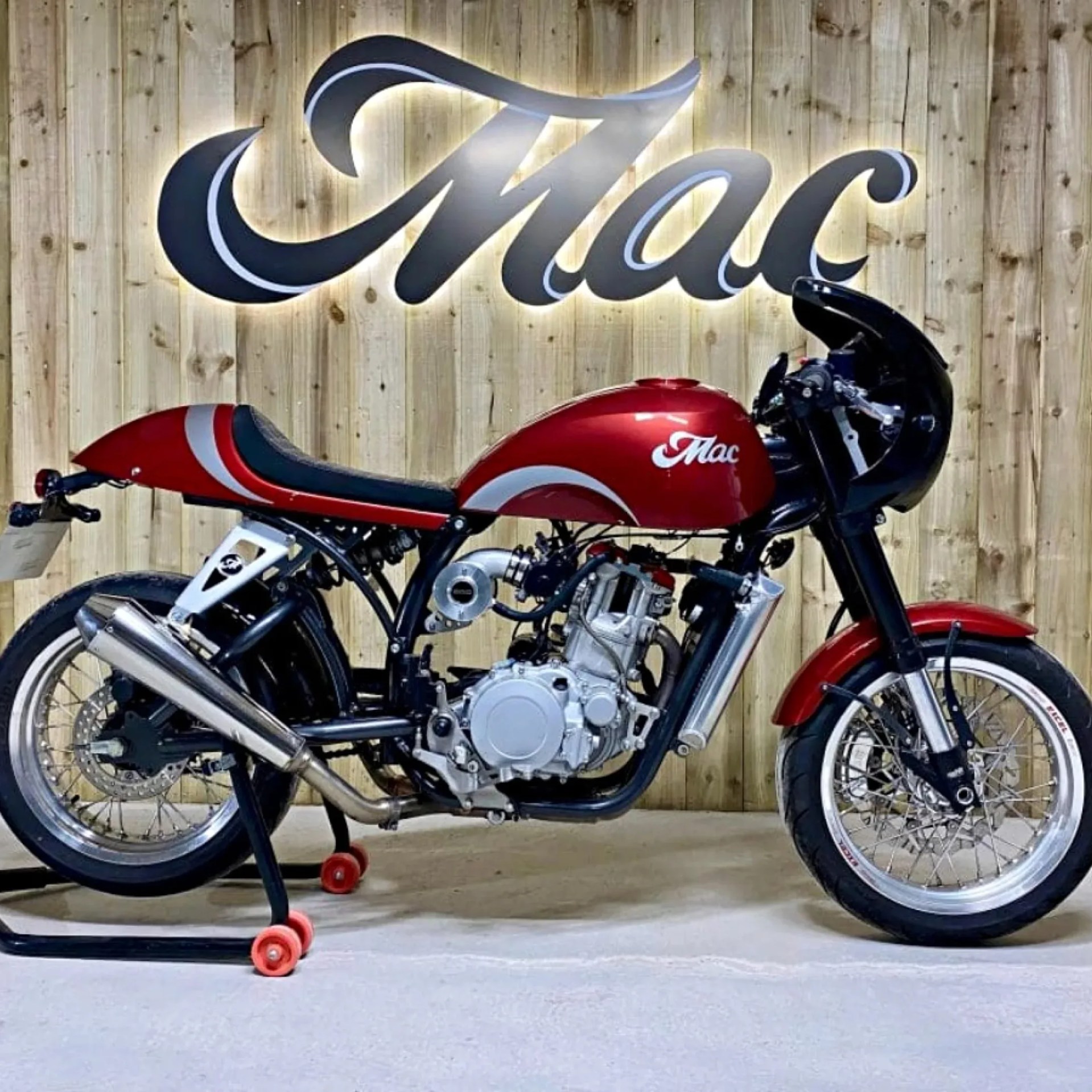 Red and black cafe racer motorcycle with white accents parked on a stand in front of a wooden wall with illuminated "Mac" sign.