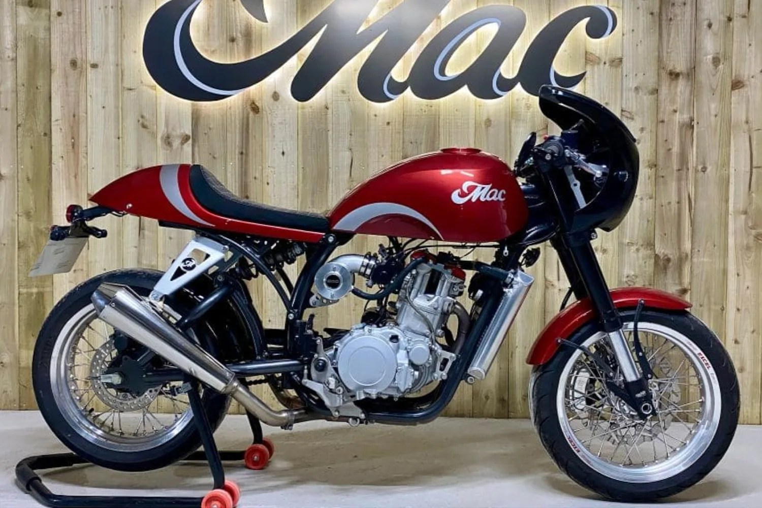 Red and black cafe racer motorcycle with silver exhaust and "Mac" logo, displayed on a stand against a wooden wall.