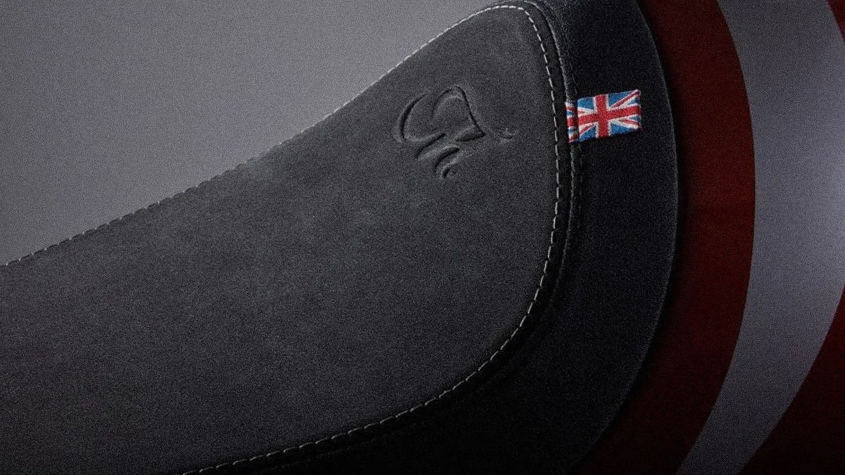 Close-up of a dark gray suede motorcycle seat with white stitching and a small Union Jack flag tag.