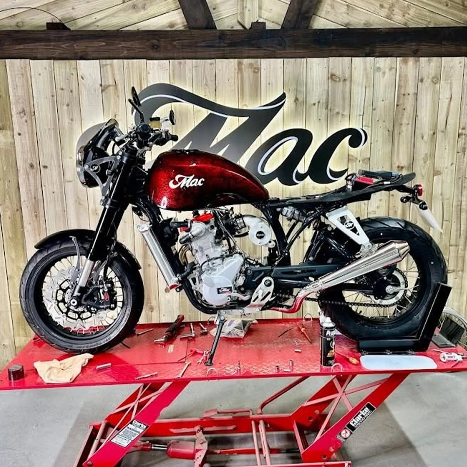 Red and black motorcycle frame with a red fuel tank labeled "Mac" on a red lift table with tools scattered around.