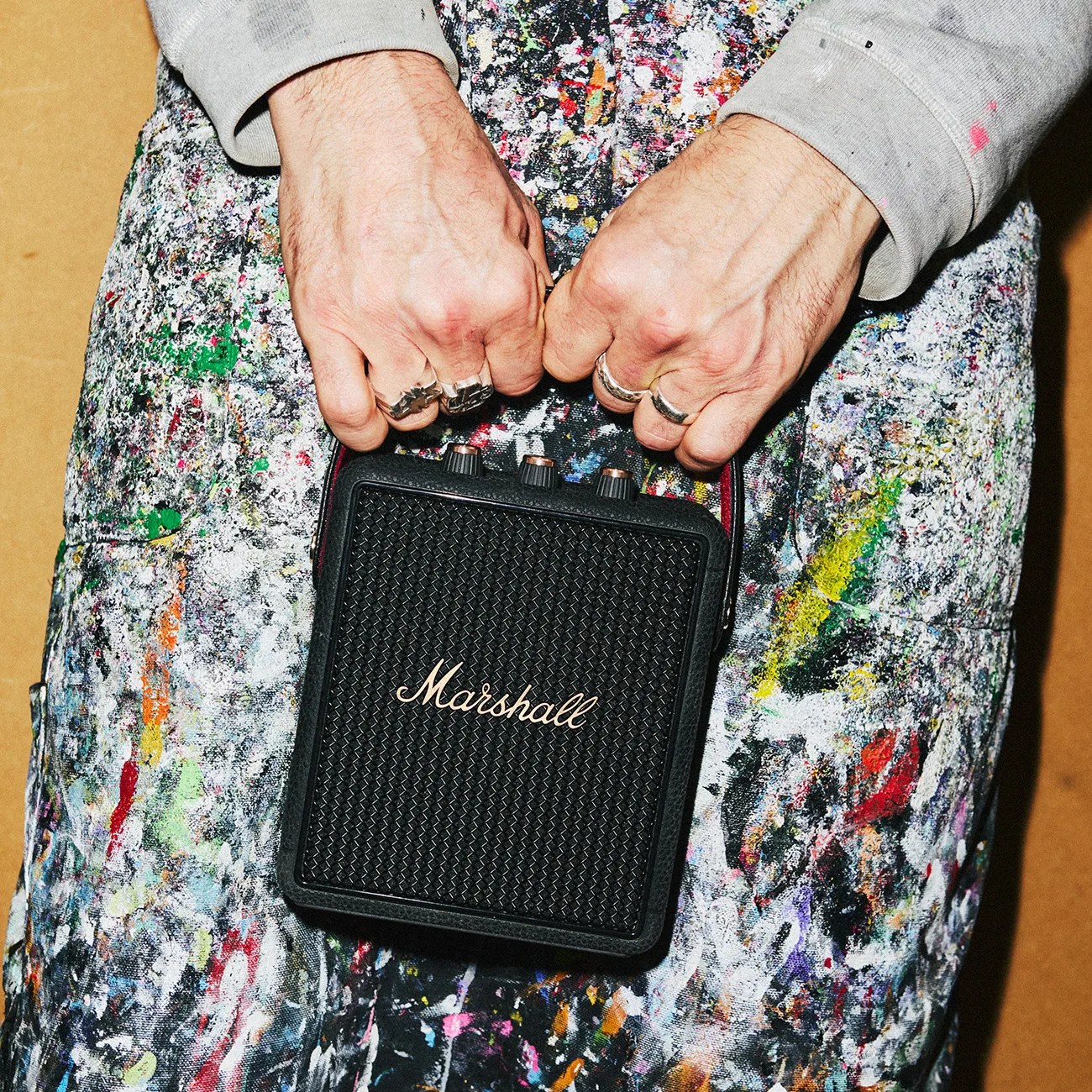Hands holding a black Marshall portable speaker against paint-splattered pants.
