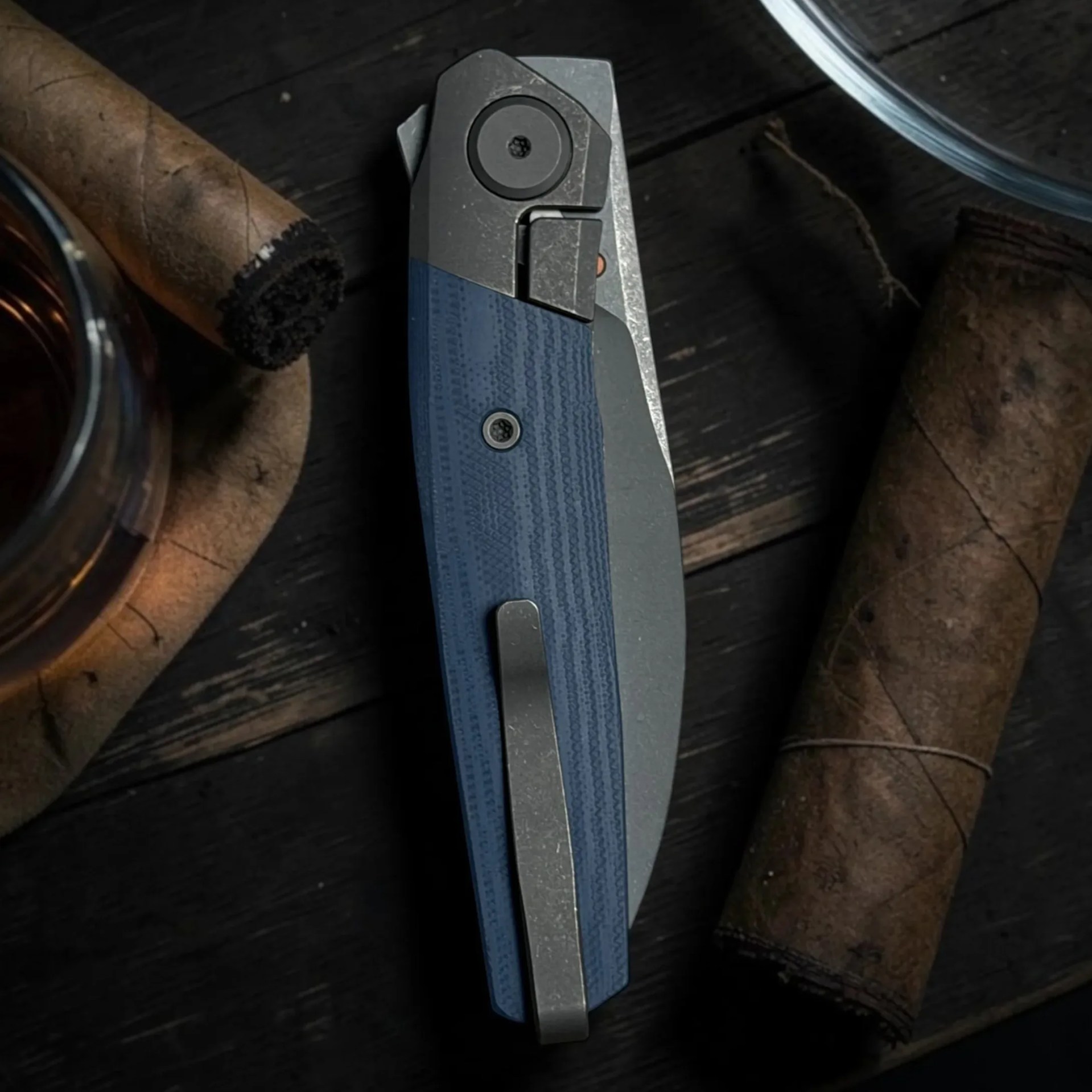Folded blue and gray pocket knife with metal clip on a dark wooden surface next to cigars and a glass.