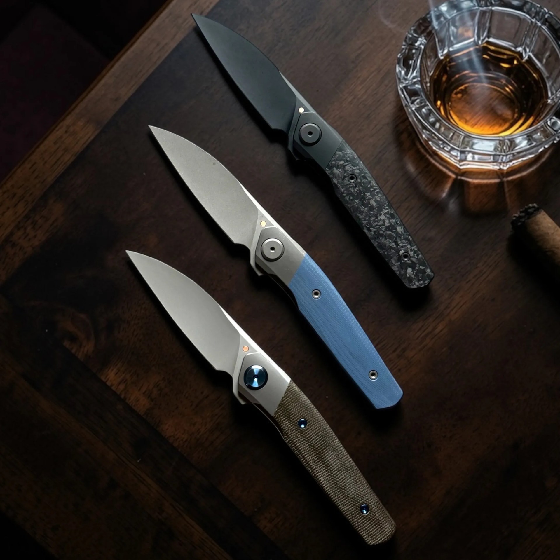 Three folding knives with different colored handles—black, blue, and tan—laid out on a dark wooden surface next to a glass of amber liquid.