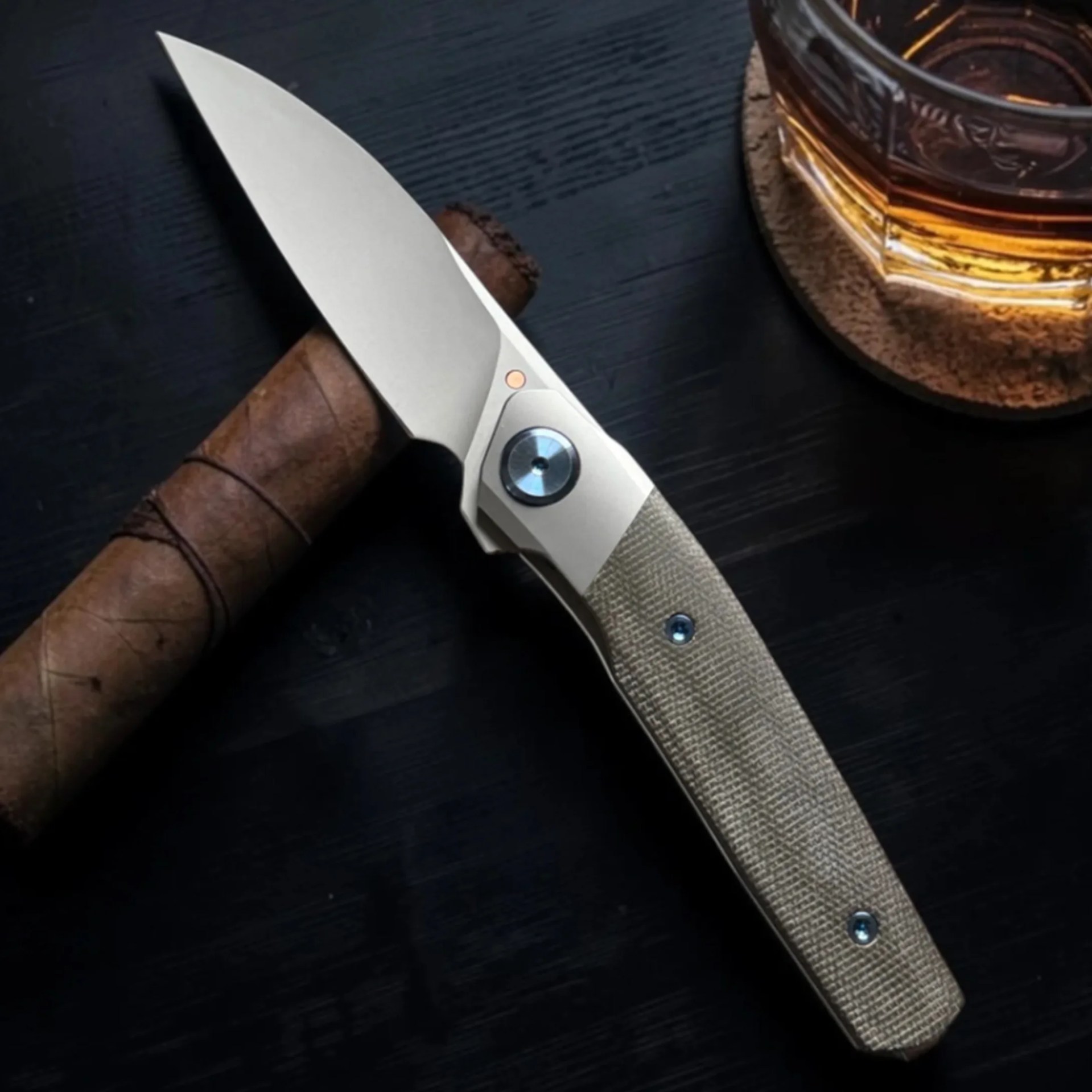 Knife with a textured beige handle resting on a cigar next to a glass of amber liquid on a dark surface.