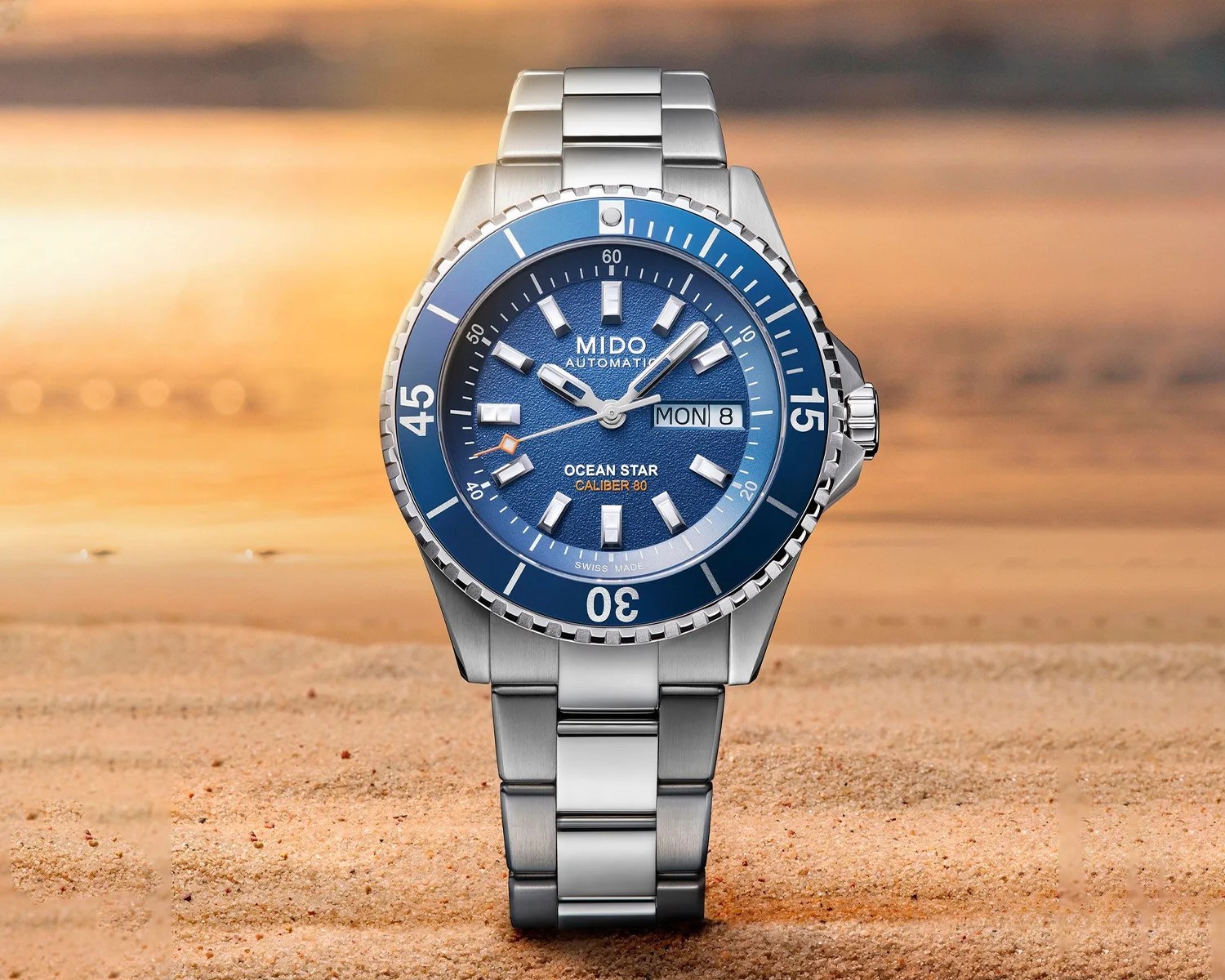 Stainless steel Mido Ocean Star watch with blue textured dial and bezel, day-date display, on sandy surface.