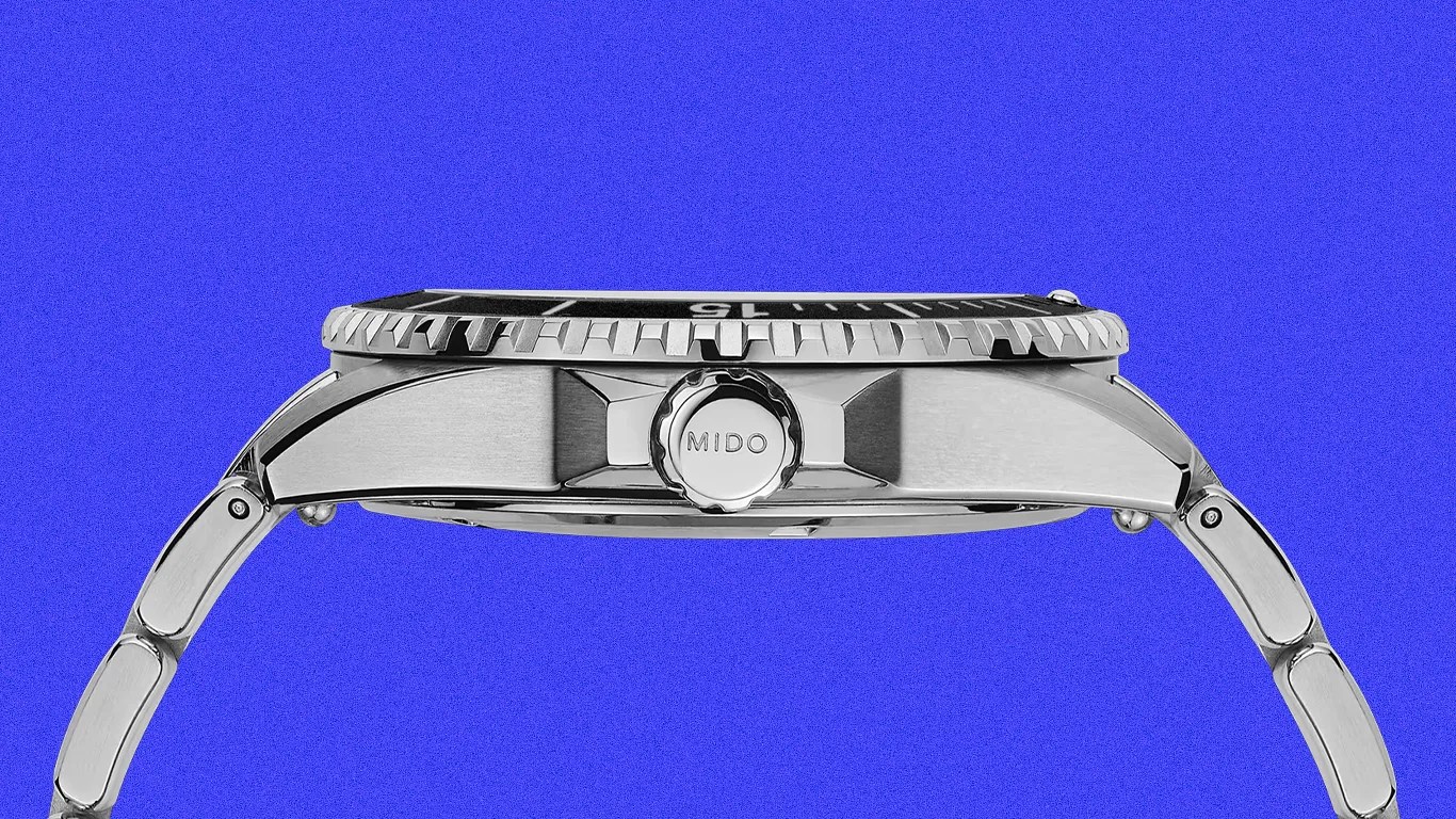Side view of a silver metal wristwatch with a ridged bezel and a crown engraved with "MIDO" against a blue background.