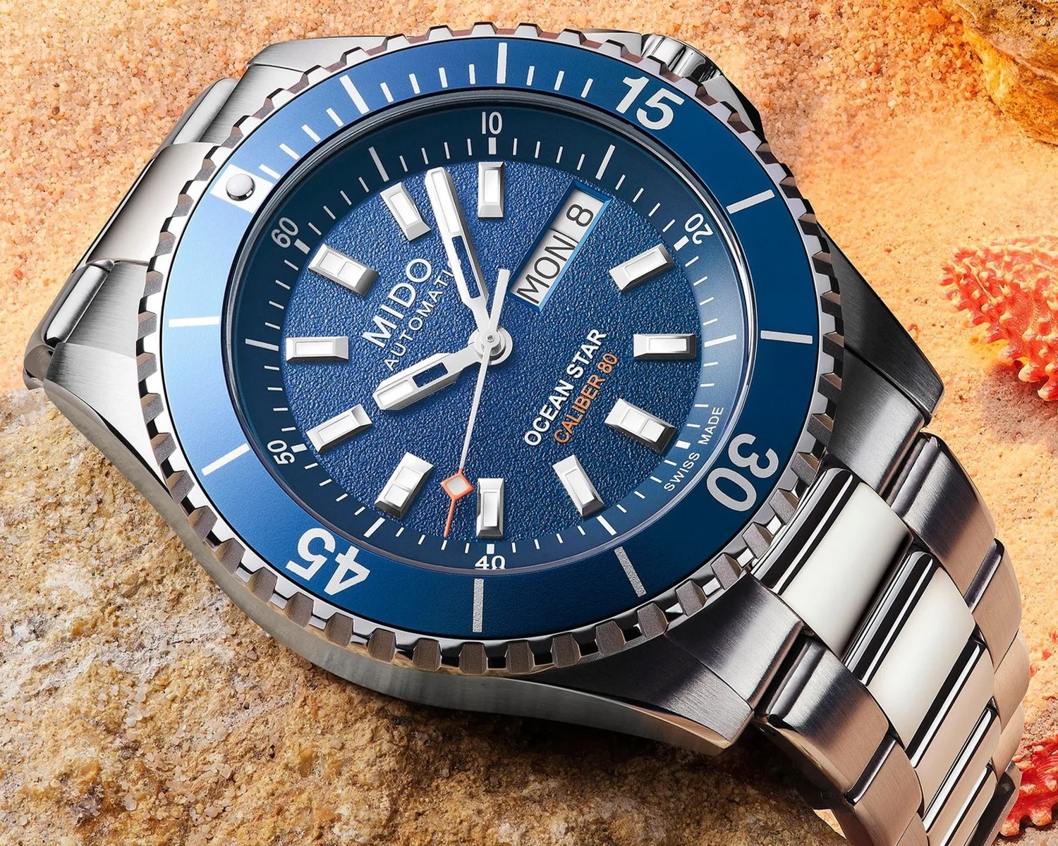 Blue dial Mido Ocean Star Caliber 80 automatic watch with stainless steel bracelet on sandy surface.
