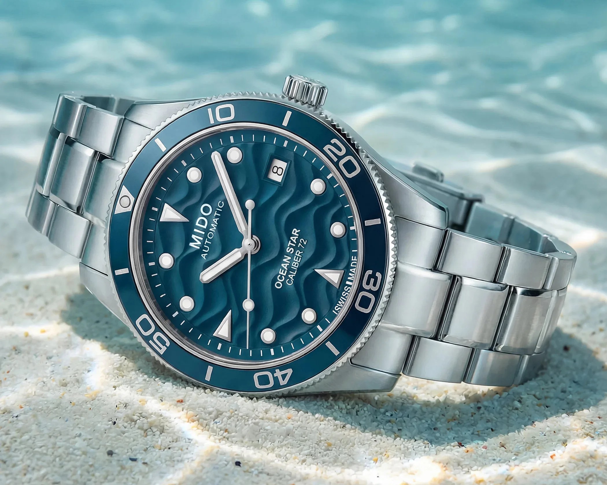 Silver metal wristwatch with a blue wave-patterned dial and rotating bezel on sandy surface.