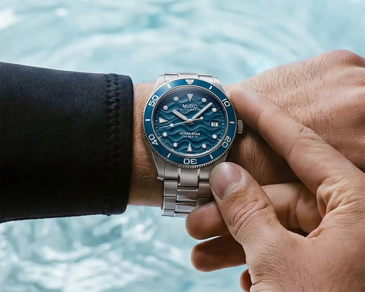 Silver metal wristwatch with a blue wave-patterned dial and bezel, worn on a wrist above water.