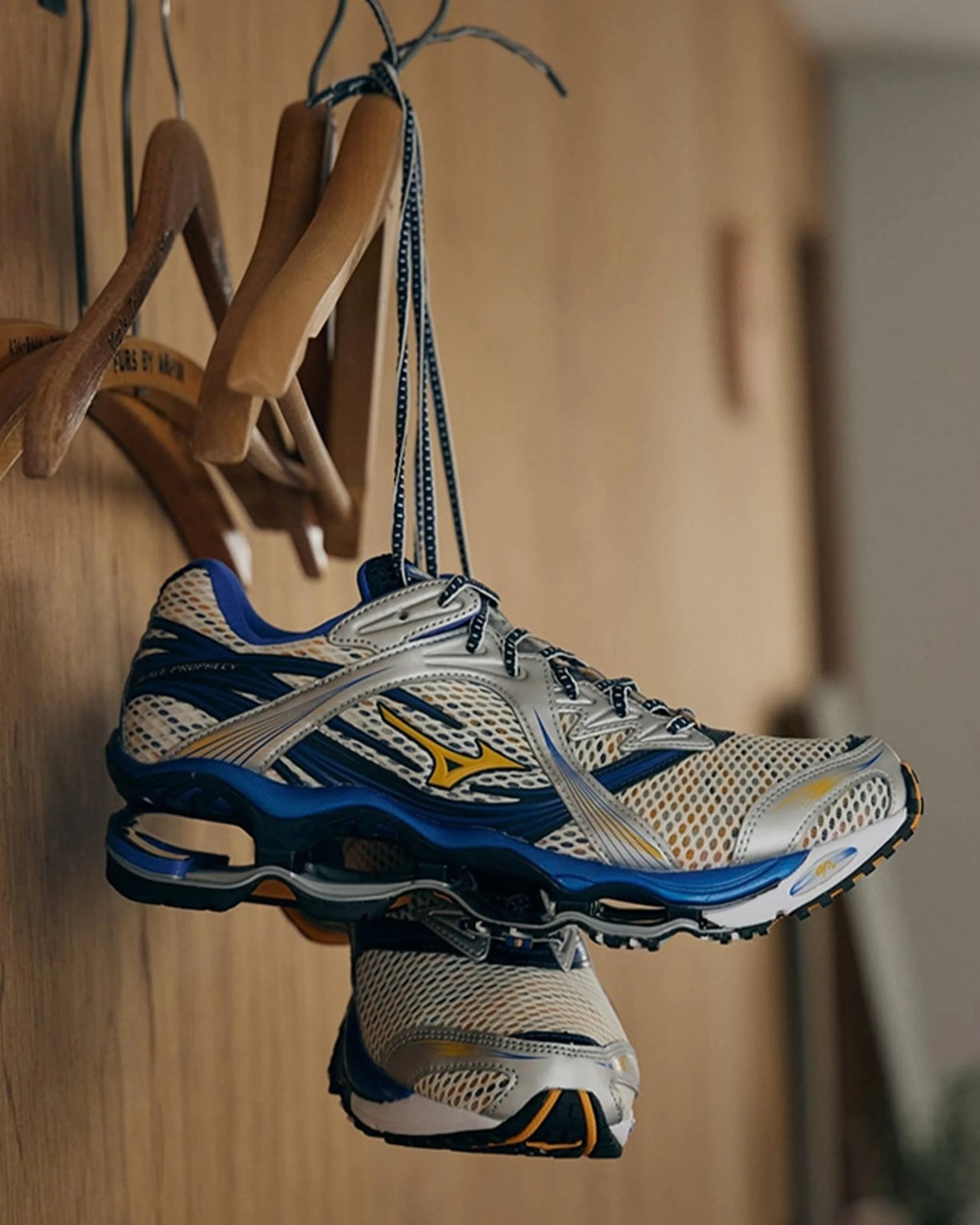 White and blue Mizuno running shoes hanging by laces on wooden hangers against a wood wall.