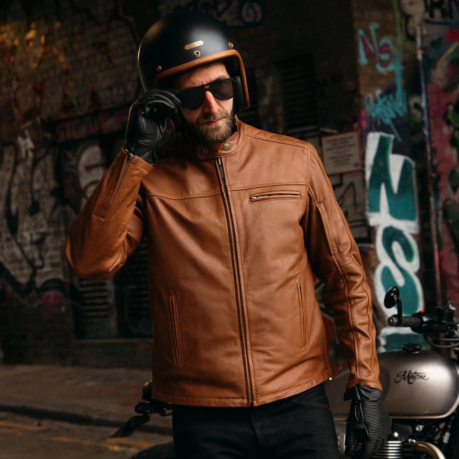 Man wearing a brown leather jacket, black gloves, black helmet, and sunglasses standing near a motorcycle with graffiti background.