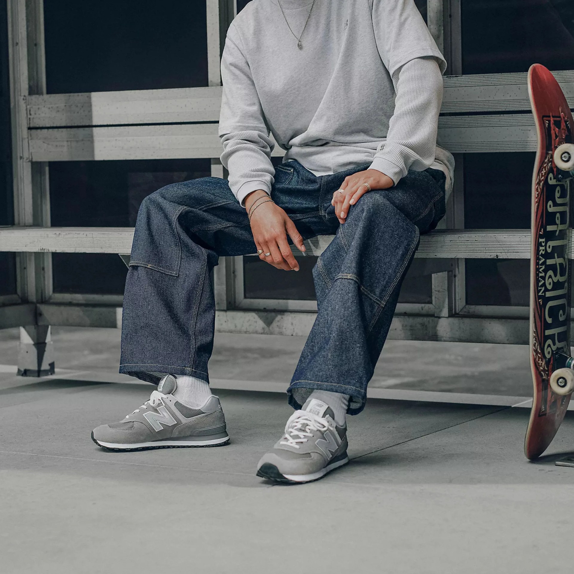 Person wearing gray layered long-sleeve shirt, dark denim pants, and gray New Balance sneakers sitting on a bench next to a skateboard.