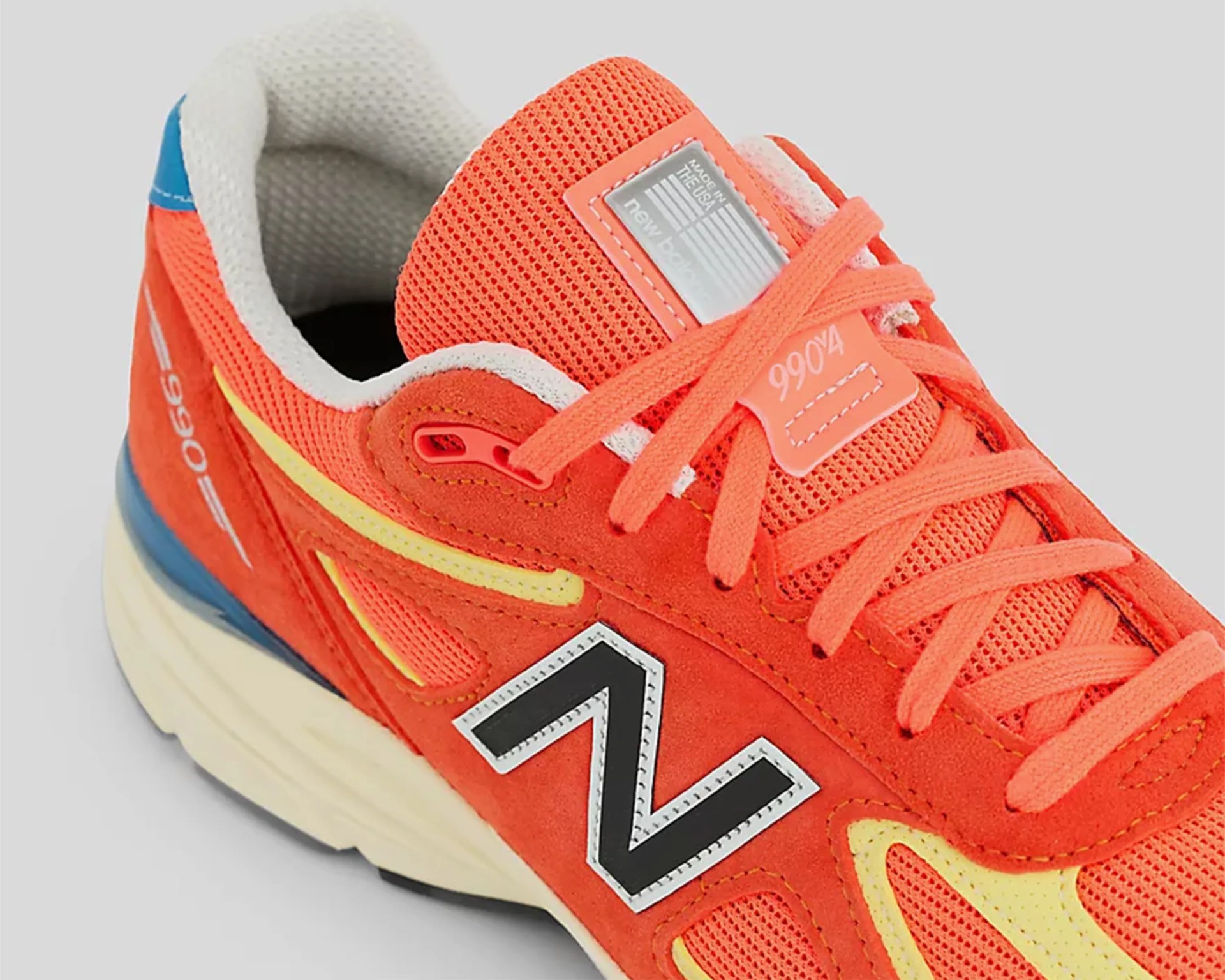 Orange New Balance 990v4 sneaker with yellow accents, black "N" logo, white sole, and blue heel detail.