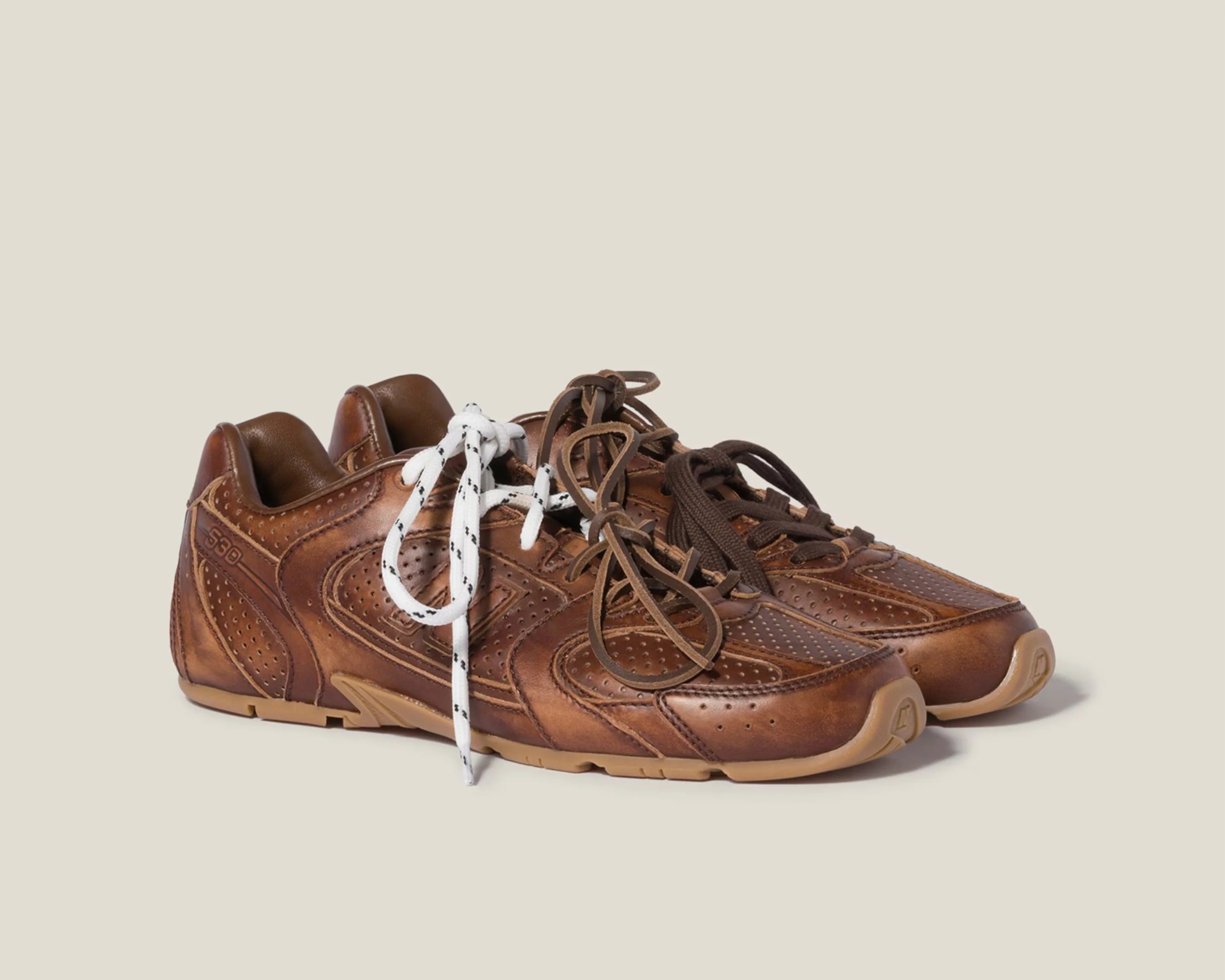 Pair of brown leather sneakers with perforated detailing and mismatched laces, one white with black dots and the other brown.