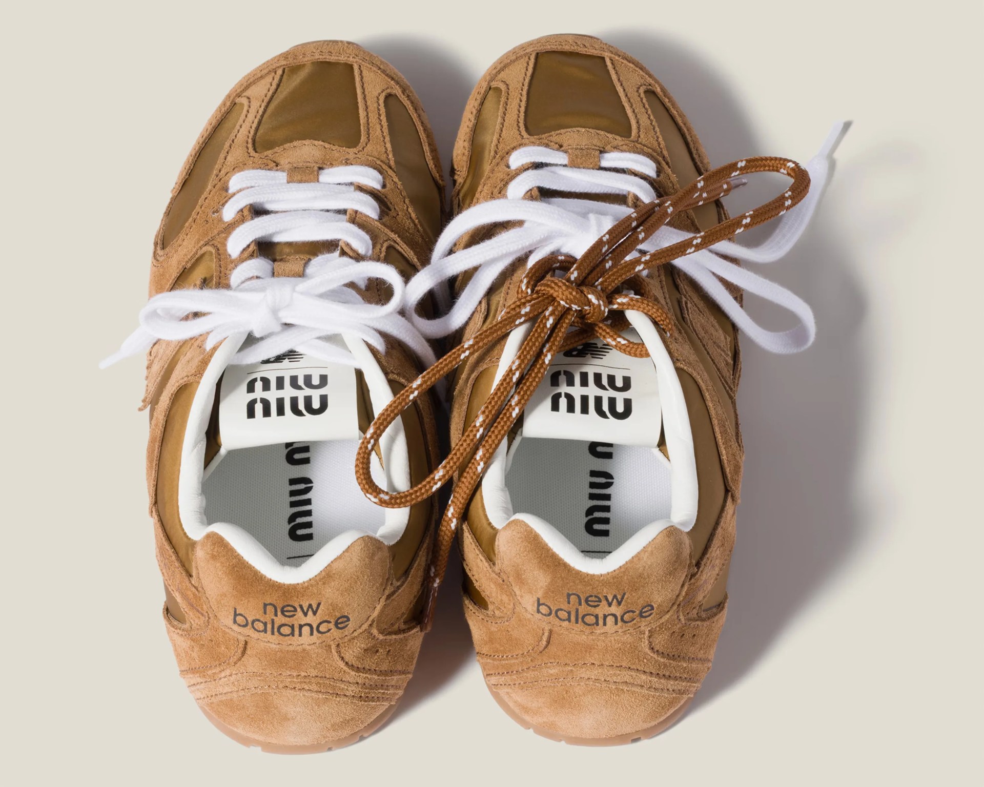 Pair of brown suede New Balance sneakers with white and brown laces on a beige background.