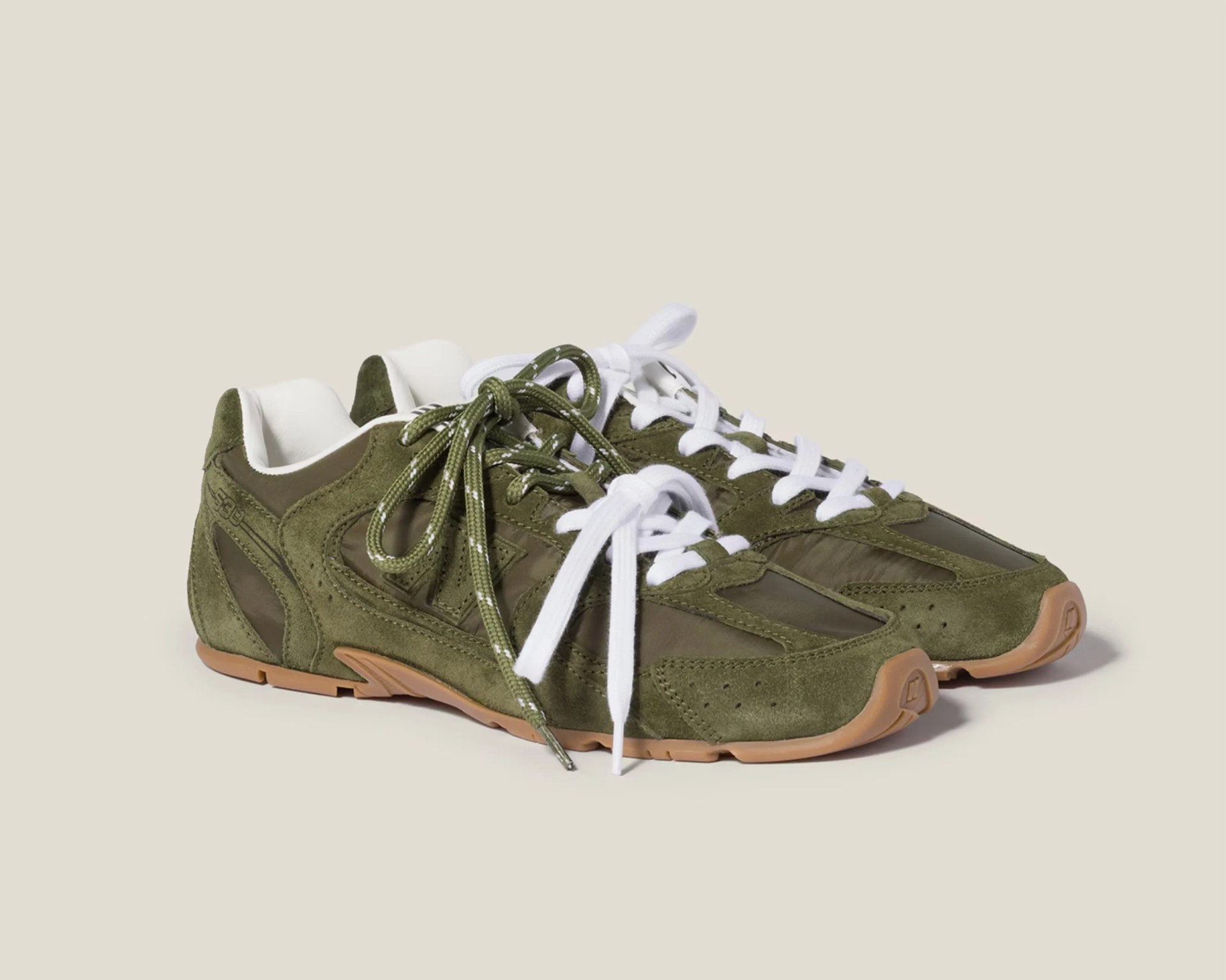 Pair of olive green suede and nylon sneakers with white and green laces and gum soles on a beige background.