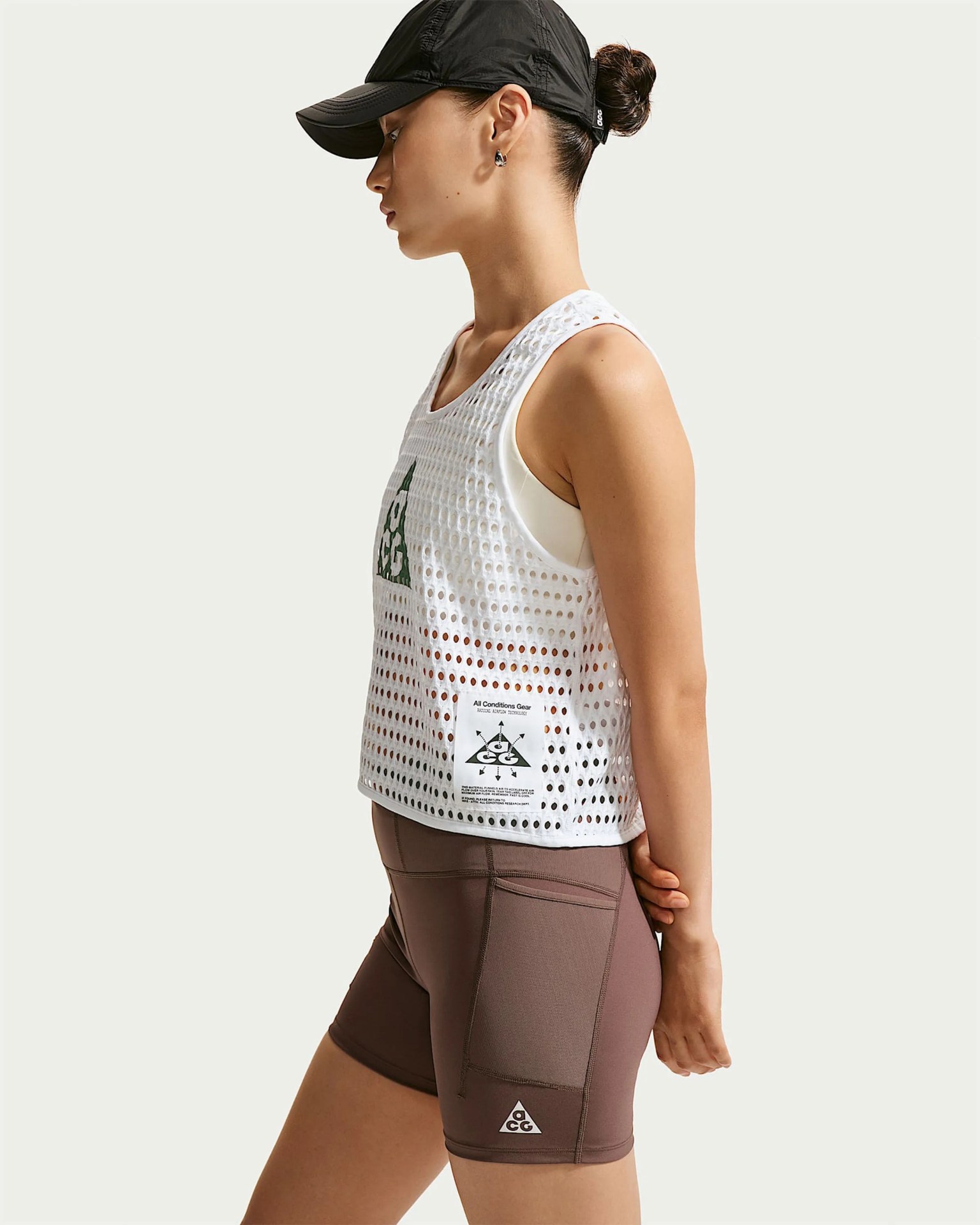 Side view of a person wearing a black cap, white mesh sleeveless top with a green logo, and brown fitted shorts with a small logo.