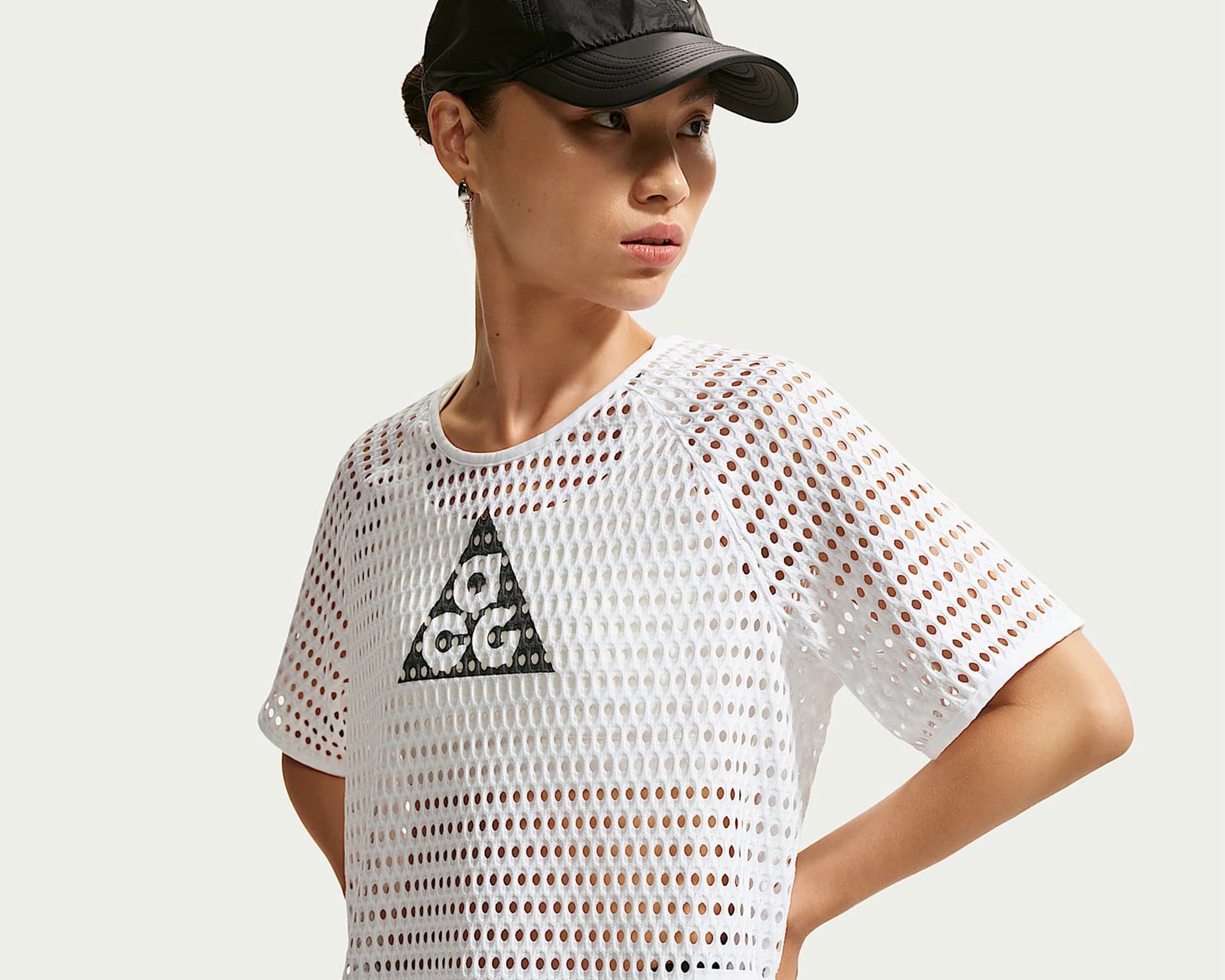 Woman wearing a white perforated mesh T-shirt with a black triangular logo and a black cap.