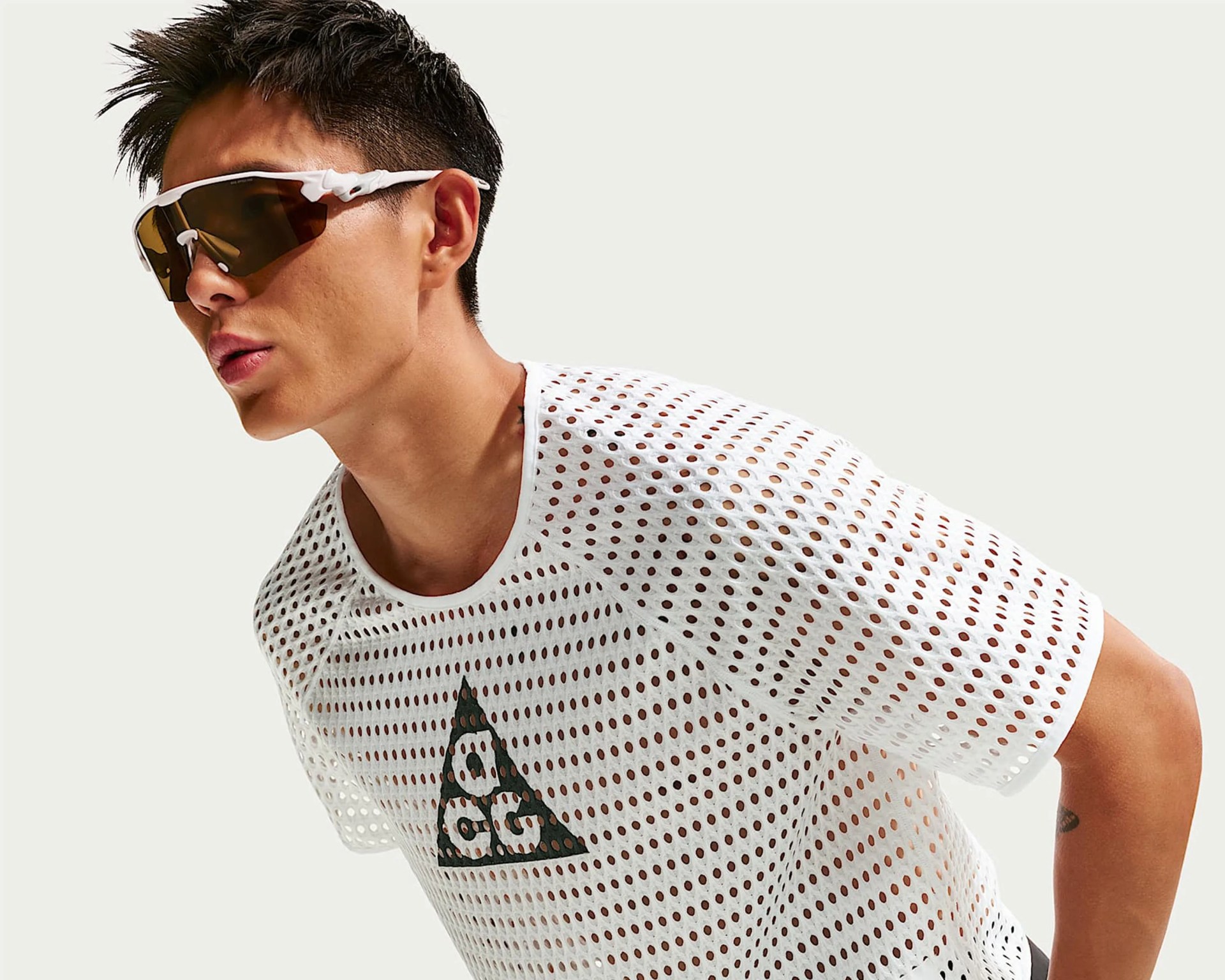 Person wearing white perforated mesh shirt with black triangular logo and white sports sunglasses with brown lenses.