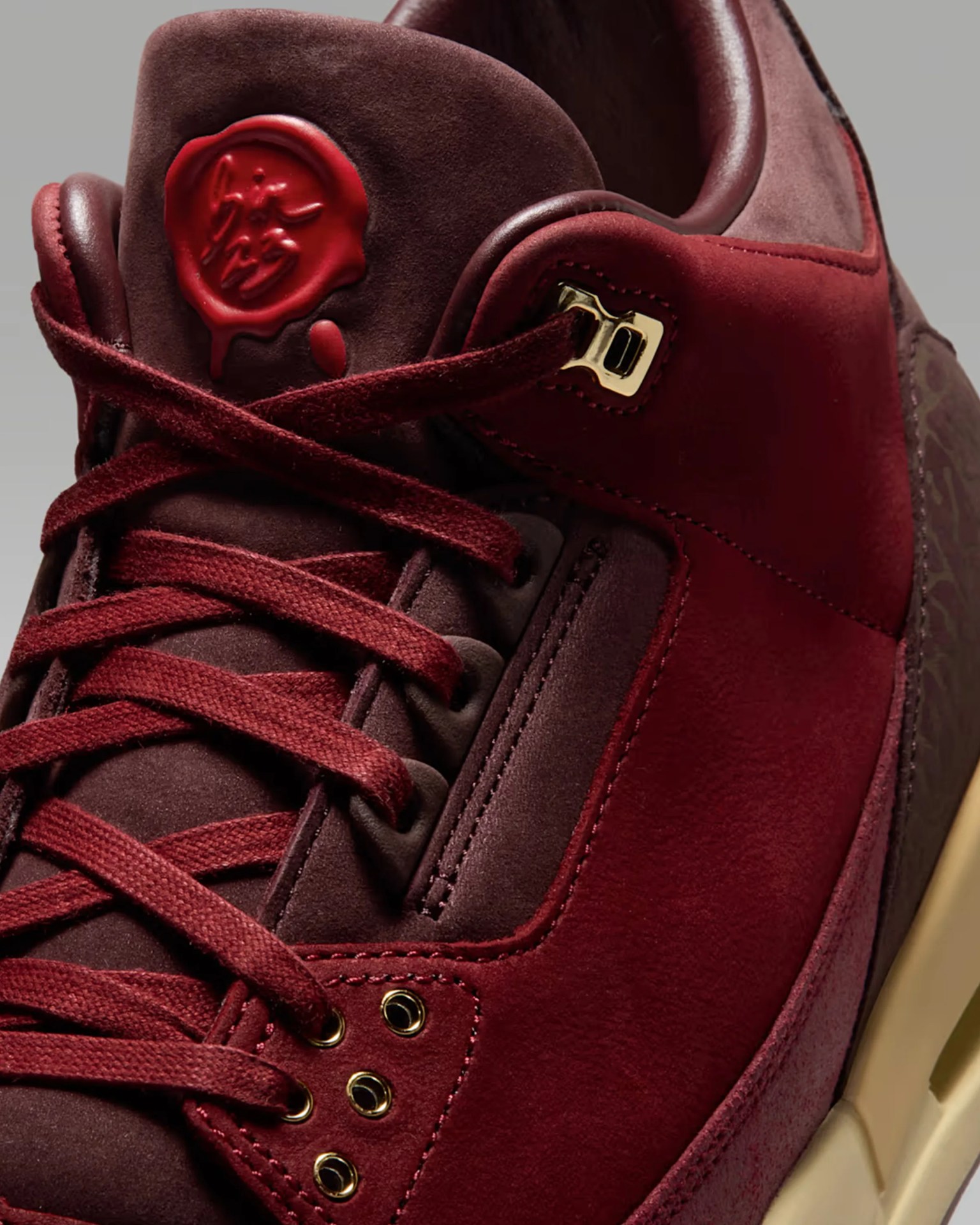 Close-up of a burgundy suede sneaker with matching laces, gold eyelets, and a red wax seal detail on the tongue.