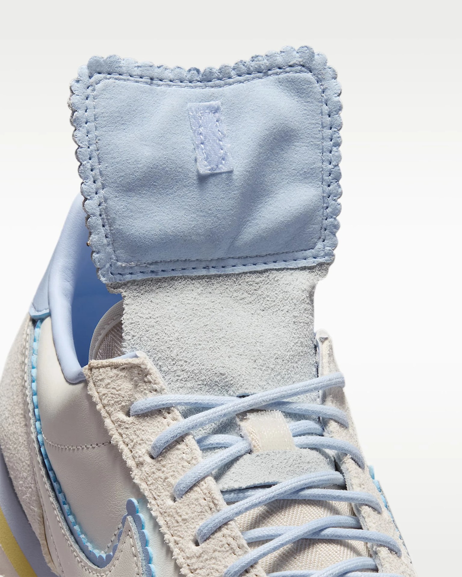 Close-up of a light blue suede tongue with scalloped edges on a beige and white sneaker with light blue laces.