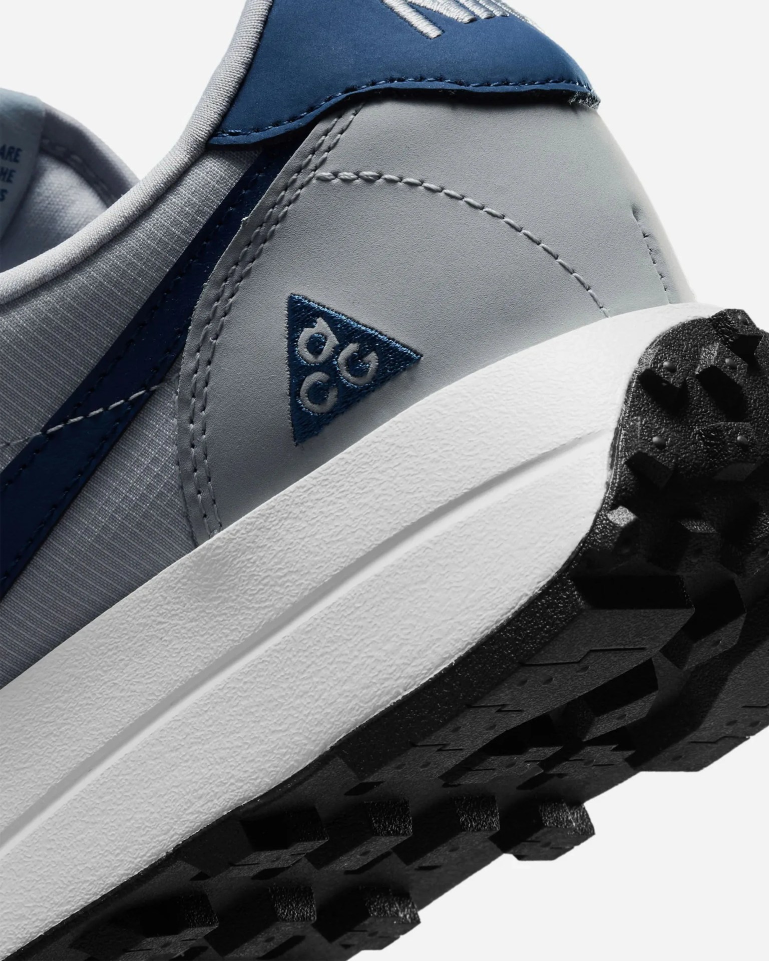 Close-up of a gray and navy sneaker heel with a white midsole and black textured outsole, featuring a blue triangular logo.