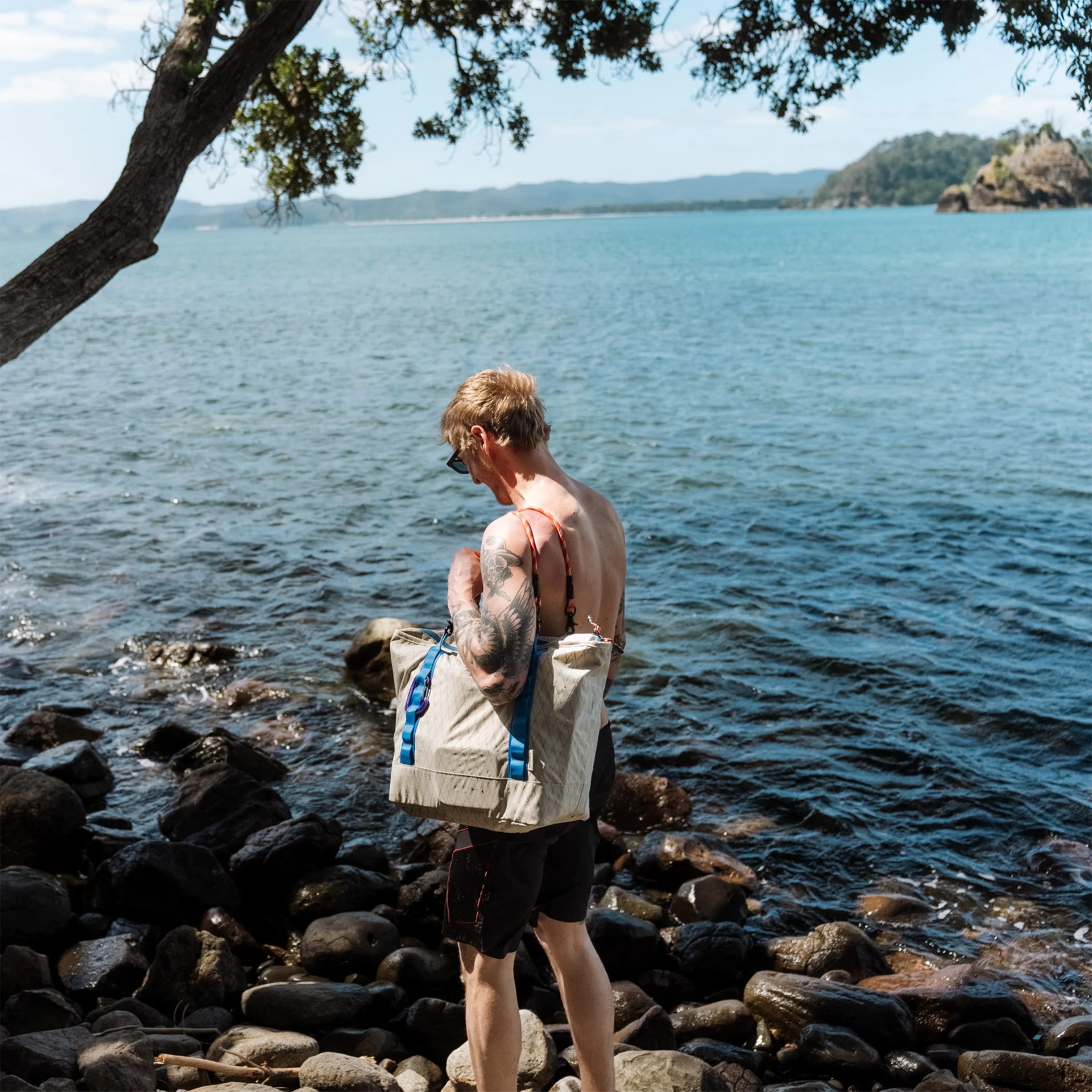 Person with tattoos wearing sunglasses and black shorts carrying a beige tote bag with blue straps by a rocky shoreline.