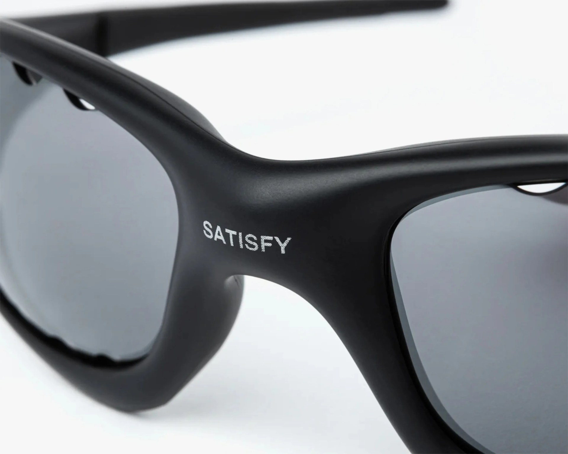 Close-up of black matte sunglasses frame with dark tinted lenses and "SATISFY" printed on the bridge.
