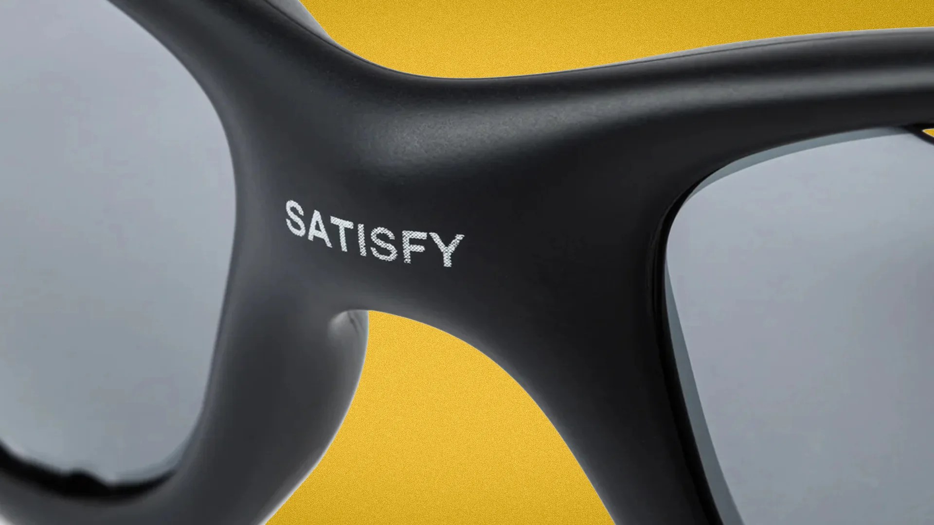 Close-up of black sunglasses frame with "SATISFY" printed in white on the bridge and gray lenses.