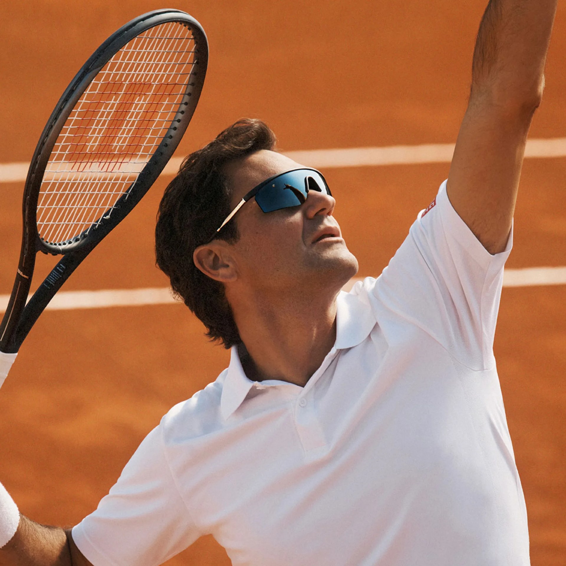 Man in white polo shirt and black sunglasses holding a tennis racket on a clay court.