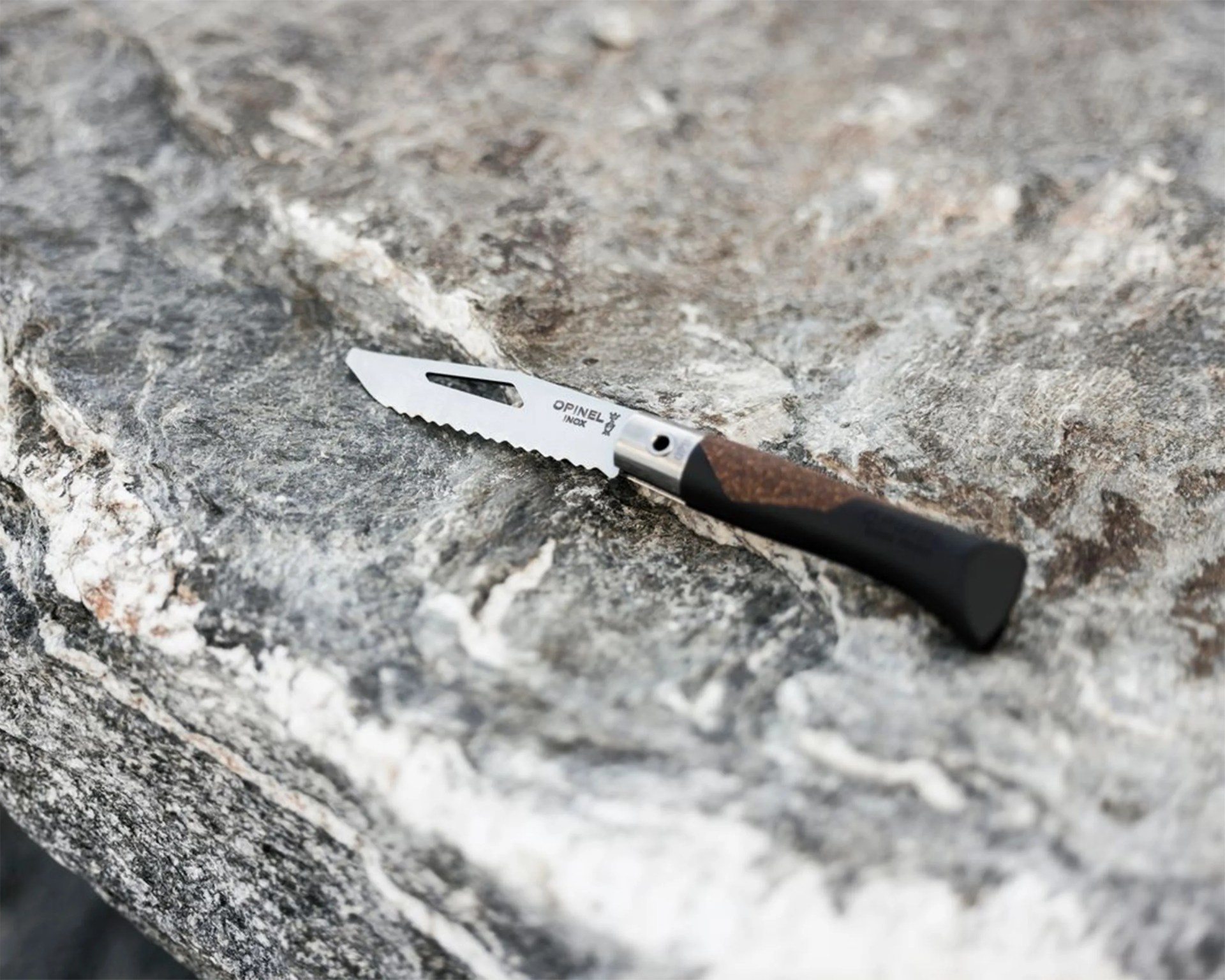 Serrated Opinel folding knife blade with wooden handle resting on a textured gray rock surface.
