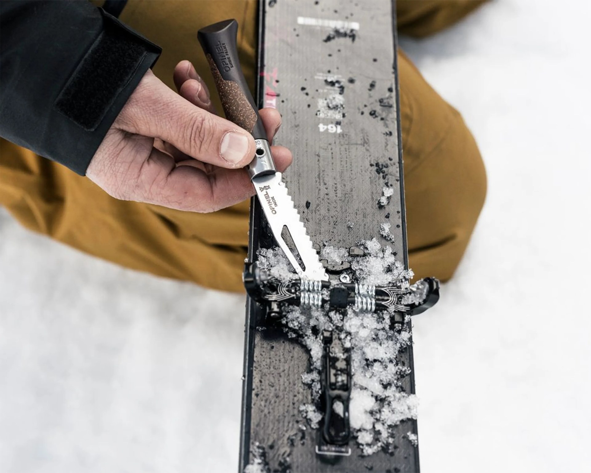 Hand holding an Opinel serrated knife scraping snow off a black ski.