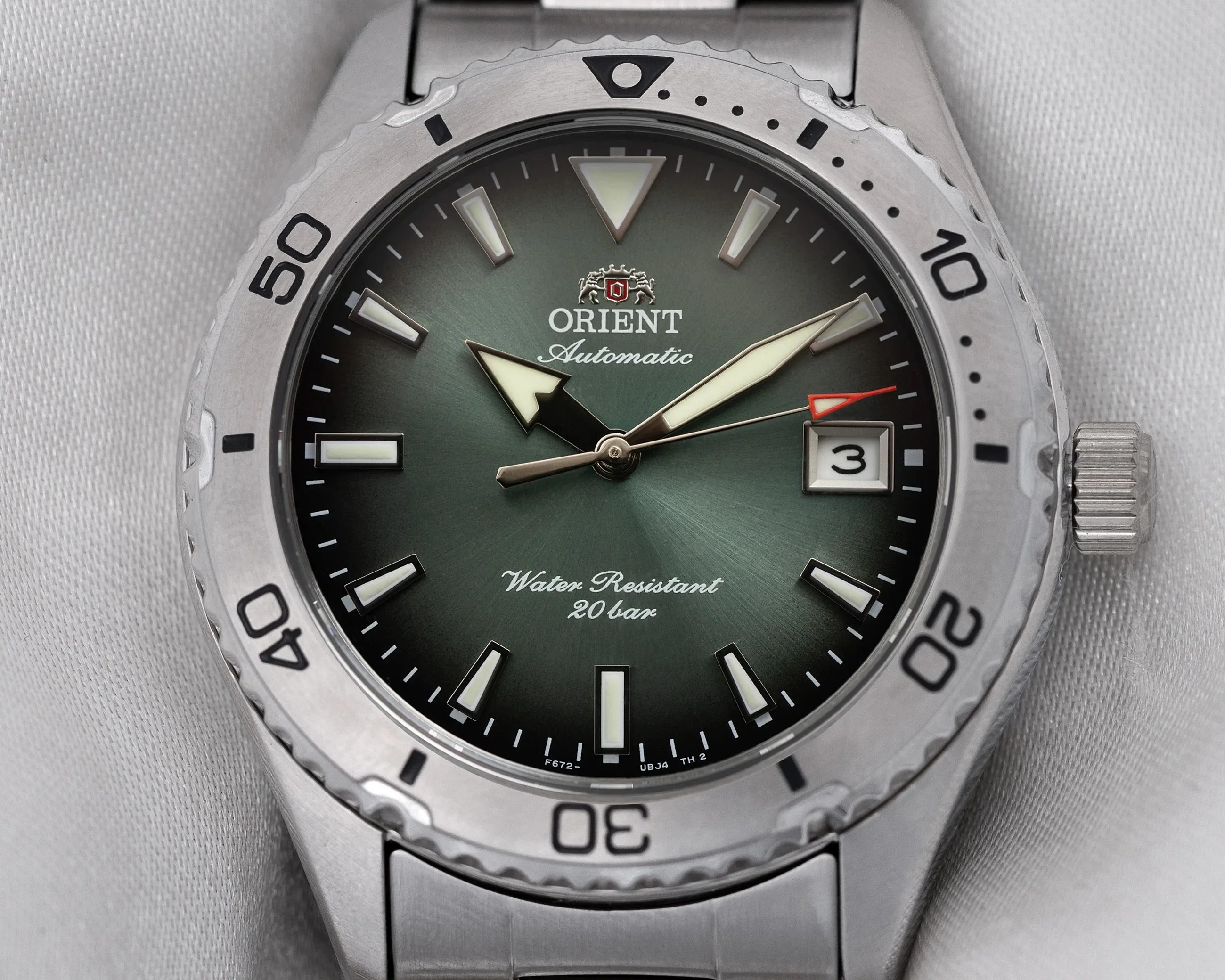 Close-up of an Orient Automatic watch with a green dial, luminous hands, date window, and stainless steel bezel and bracelet.