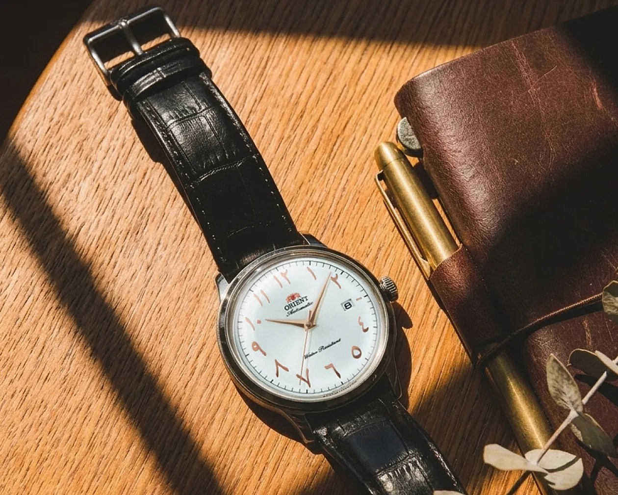 Silver Orient wristwatch with white dial and Arabic numerals on a black leather strap, next to a brown leather notebook and brass pen.